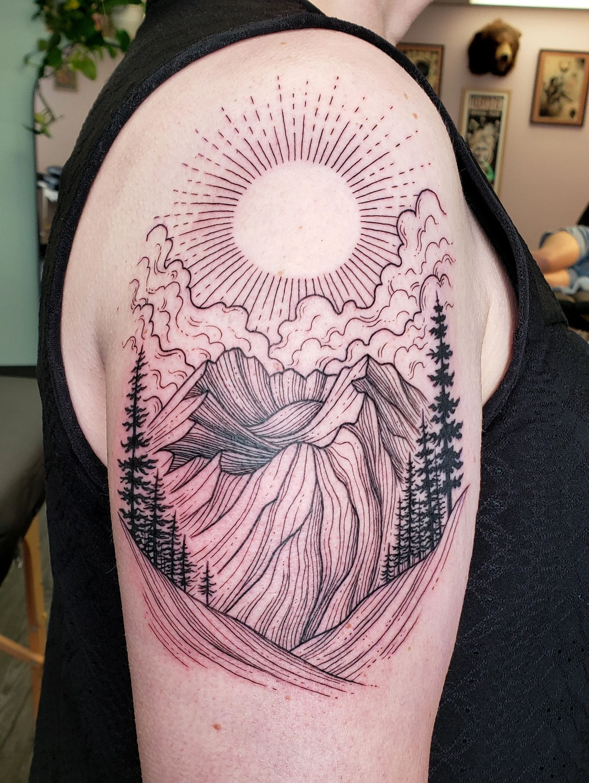 A tattoo of a sun with rays, clouds, mountains, and pine trees on a person's upper arm.