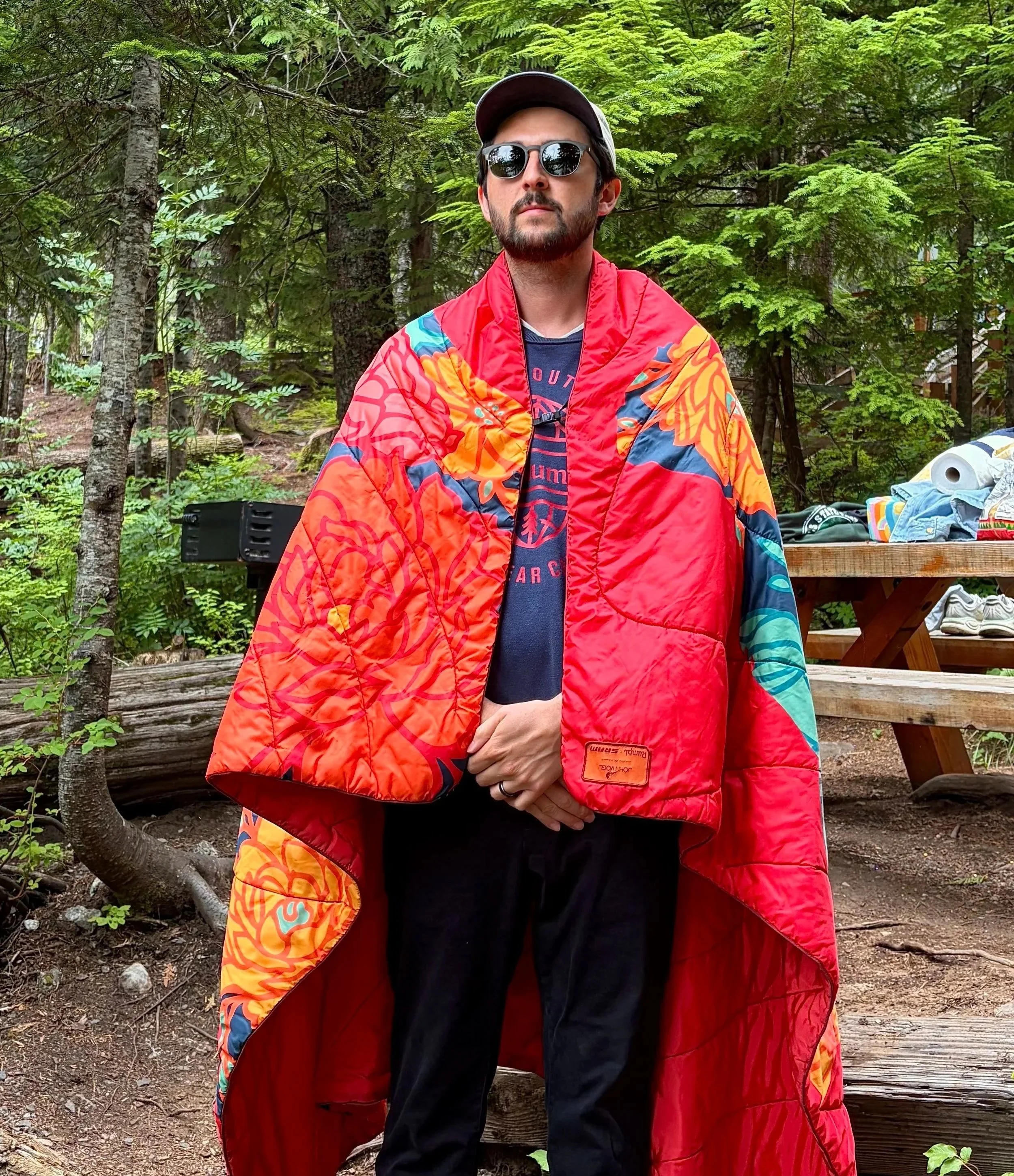 A man with a beard and sunglasses standing outdoors in a forest. He is wearing a colorful red, orange, and blue patterned jacket and a dark shirt underneath. There are trees and green foliage in the background, with some outdoor items like a grill and clothes visible on a wooden table nearby.