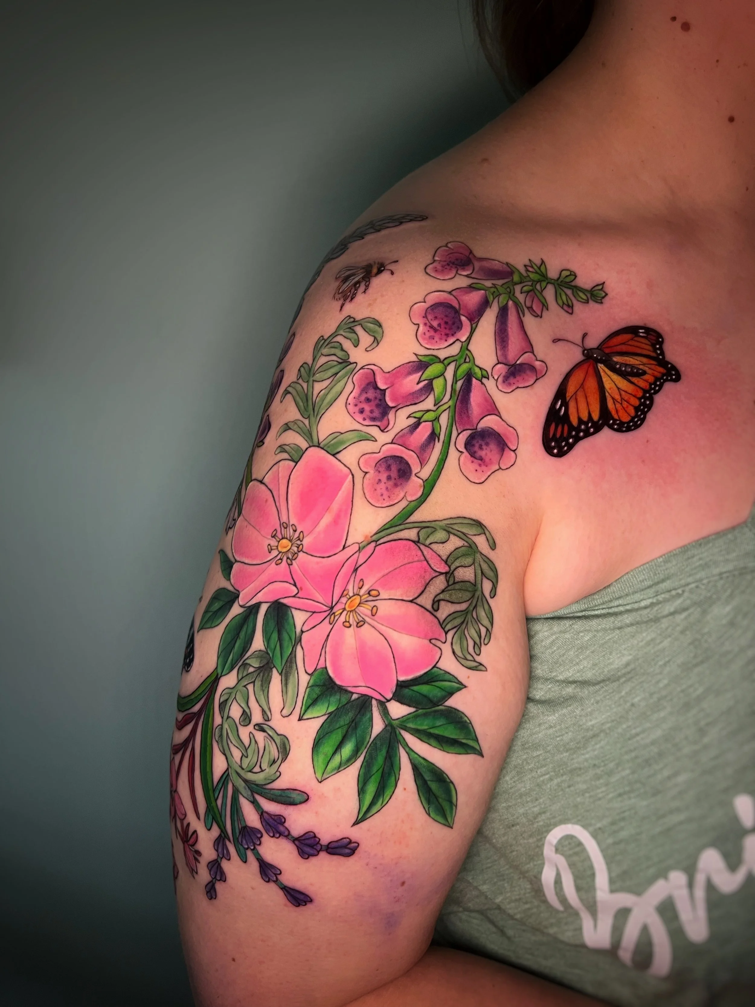 Colorful tattoo on a person's shoulder and upper arm featuring pink flowers, purple flowers, a butterfly, and a bee against a greenish background.