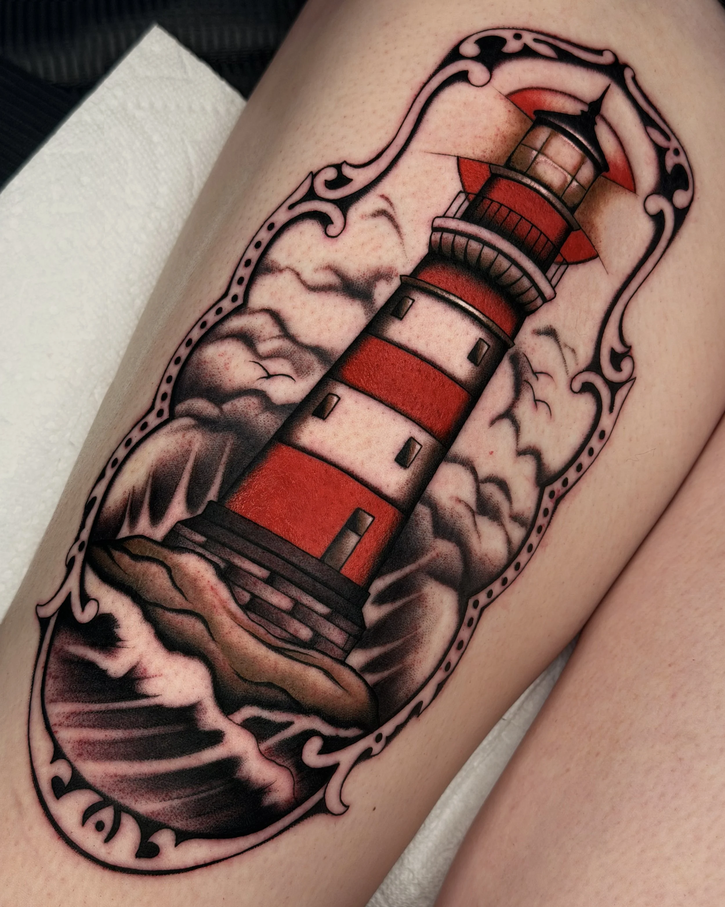 Tattoo of a lighthouse surrounded by clouds and decorative border on a person's arm