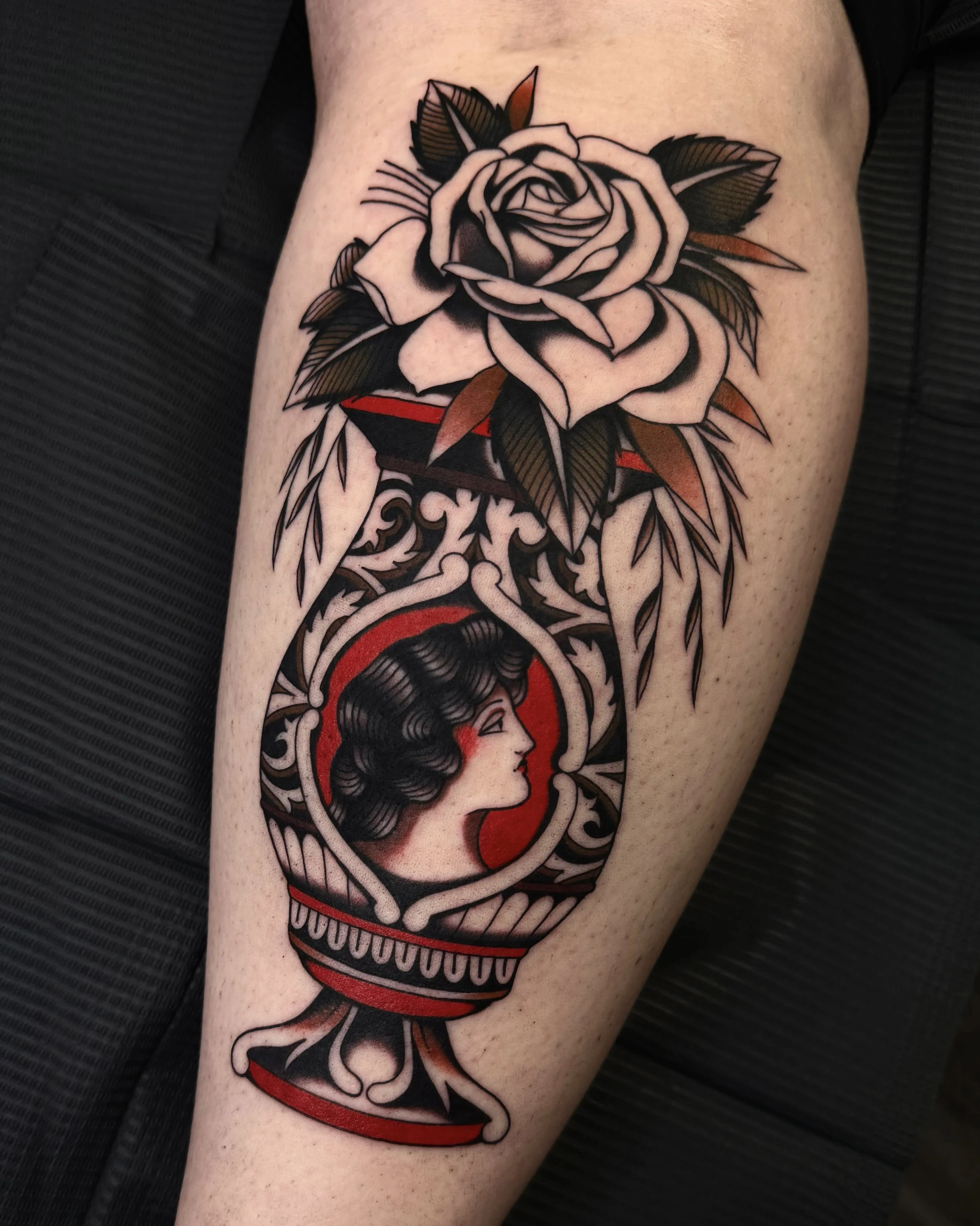 A tattoo of a woman in profile with black hair and red lips, in a traditional style, surrounded by a red and black circular frame. Above the tattoo is a black and beige rose with green leaves.