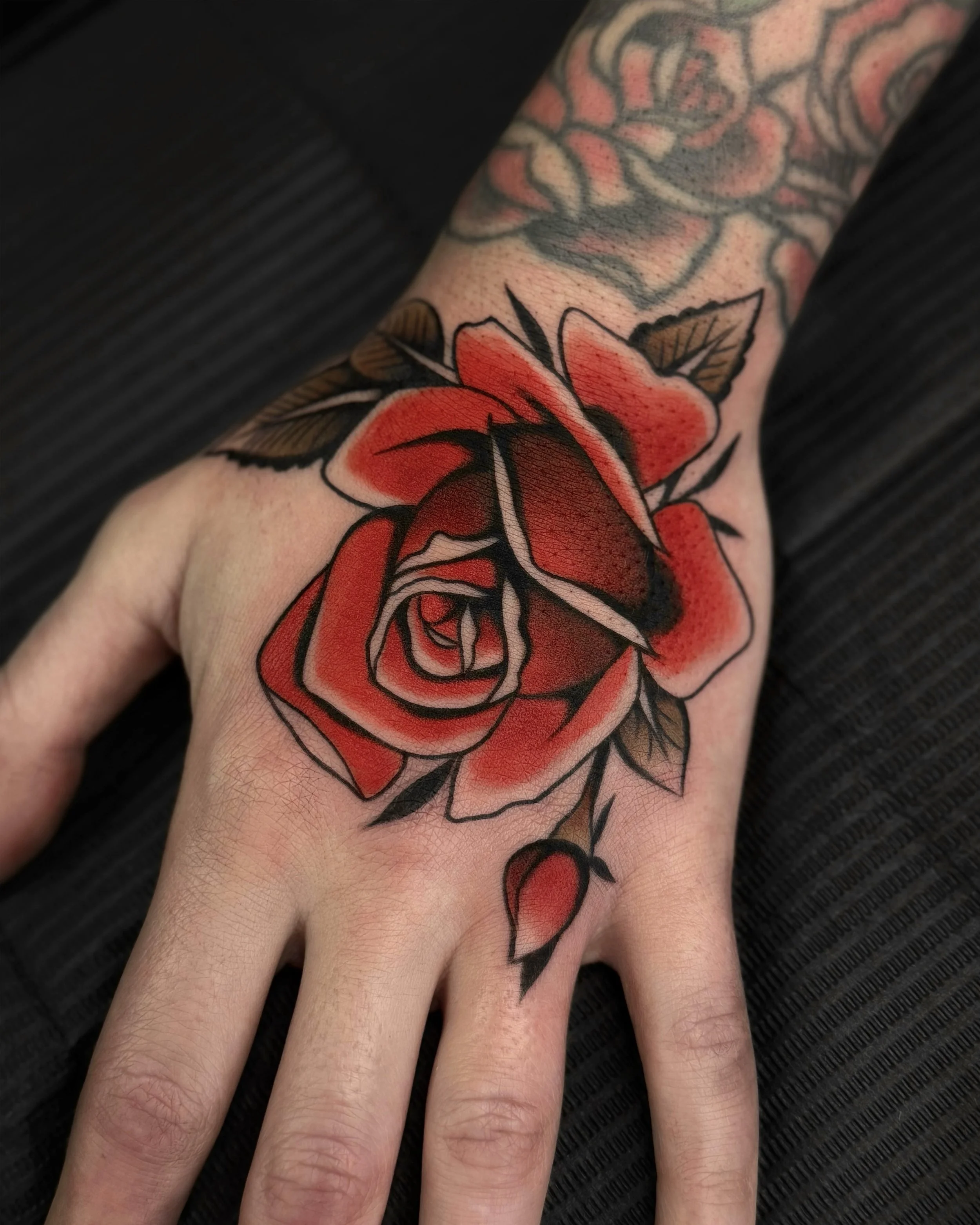 Colorful tattoo of a red and orange rose on the person's hand with black outlines and green leaves, with another tattoo visible in the background.