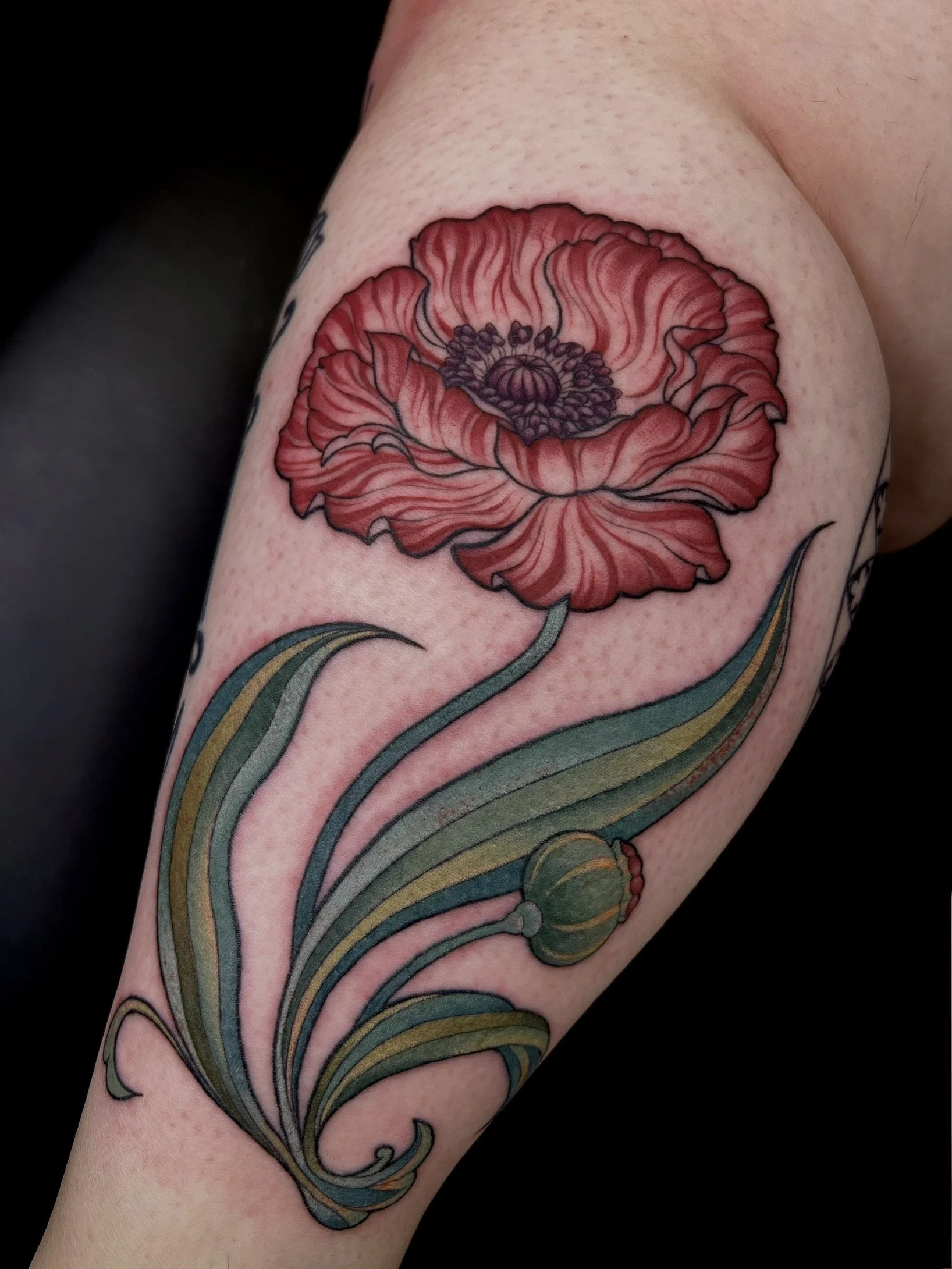Tattoo of a red poppy flower on person's upper arm with green leaves and stem, black outline, detailed shading, and vibrant colors.