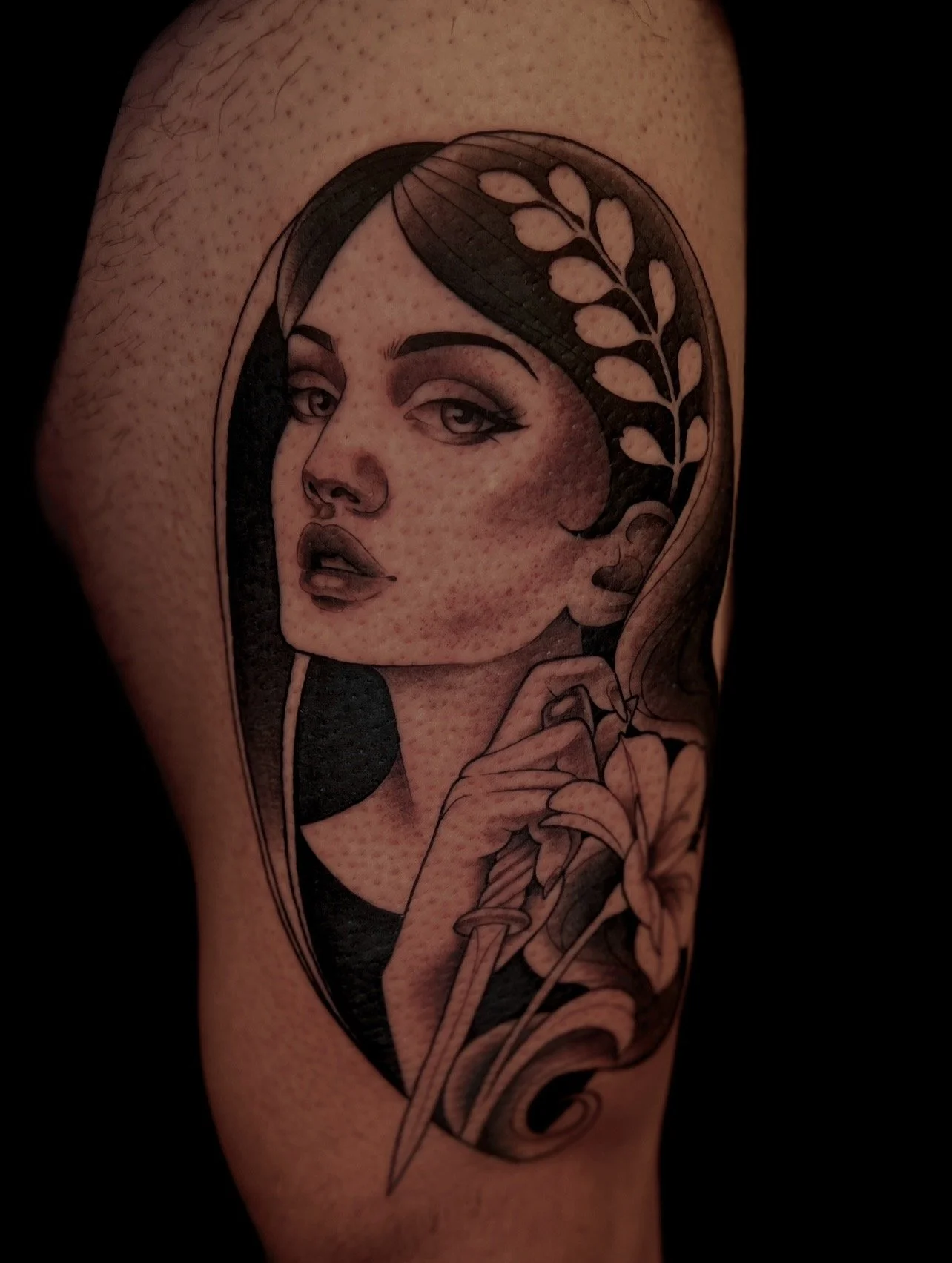 Tattoo of a woman with dark hair, makeup, and a thoughtful expression, holding a flower, with a leafy pattern on her hair, in black and gray style.