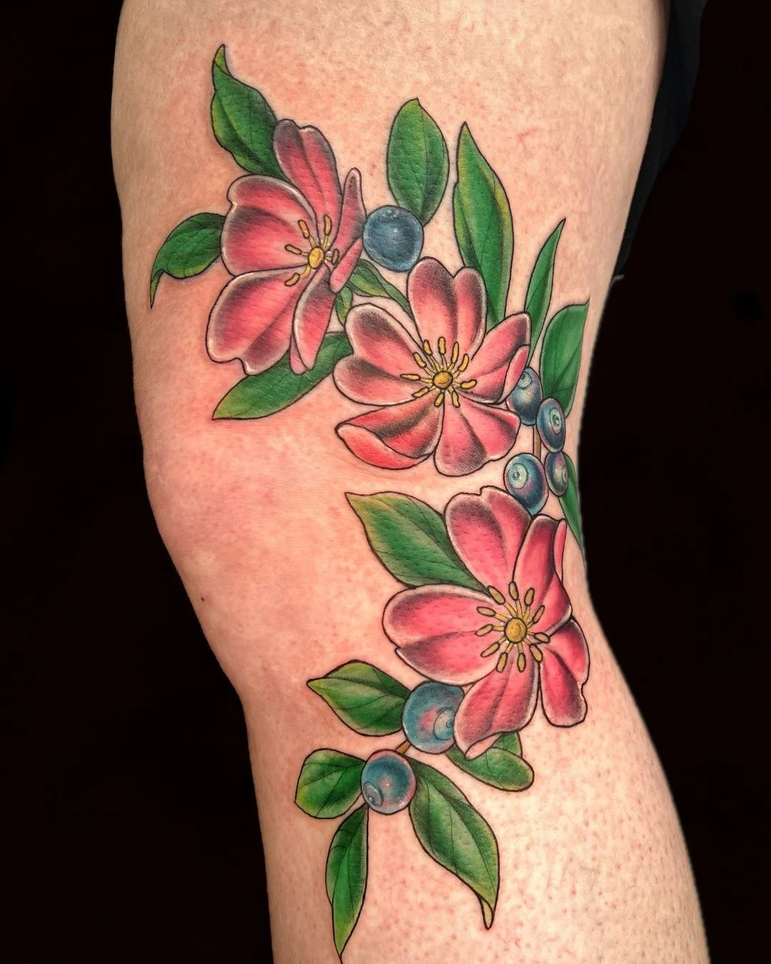 Colorful floral tattoo featuring pink flowers with yellow stamens, green leaves, and blue berries on the person's arm.