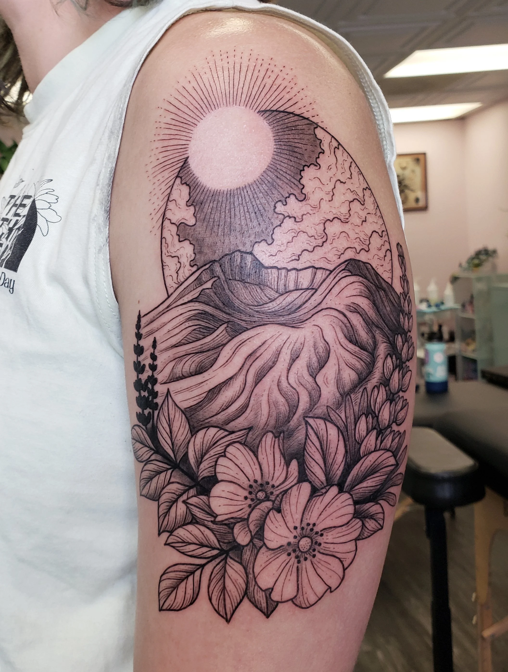 Black and pink floral tattoo on upper arm featuring a sun with rays, mountains, clouds, and blooming flowers.