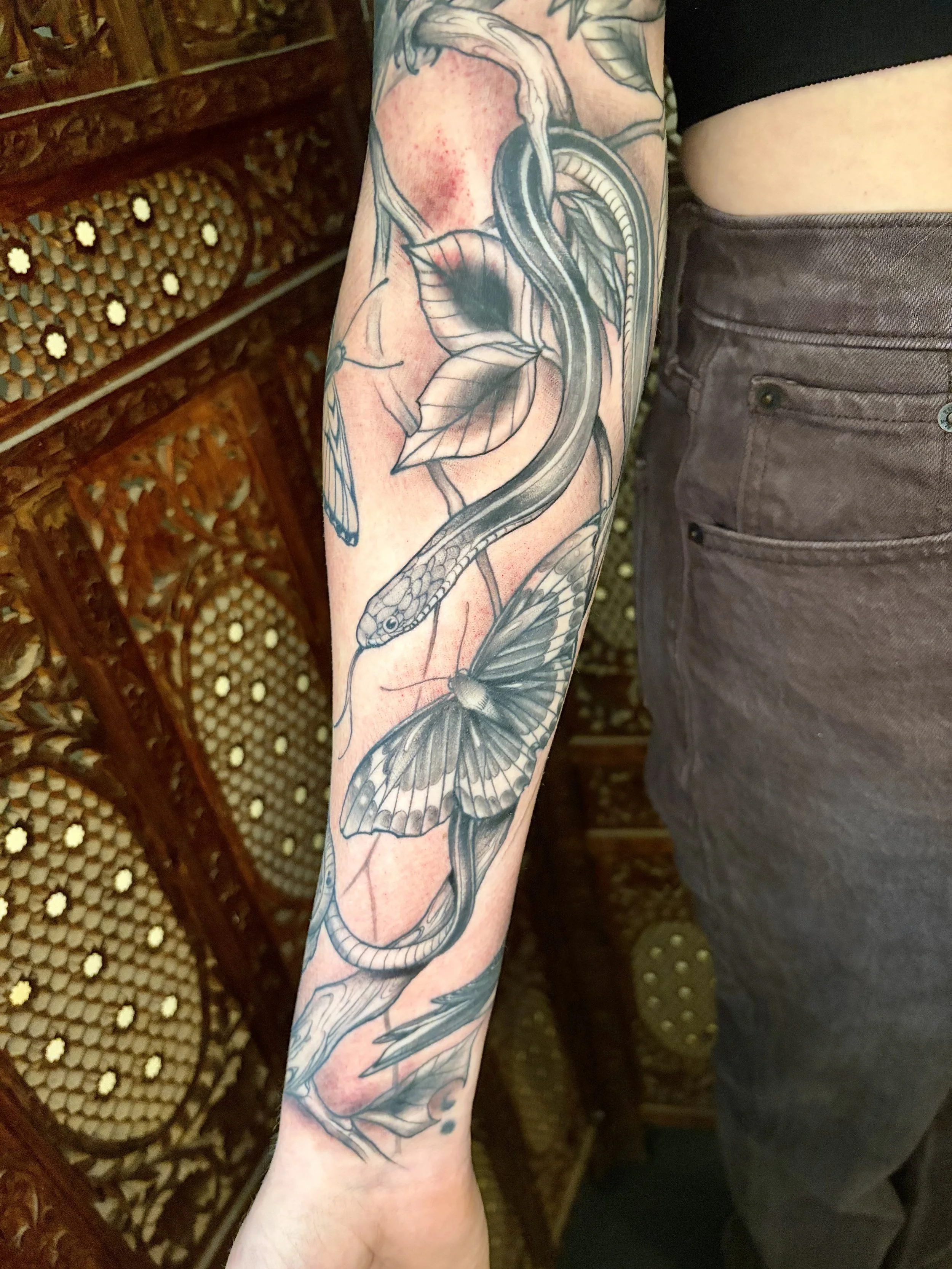 A person's arm with a tattoo of a snake weaving through leaves and a butterfly on the forearm, with a blurry background of rattan furniture and a person in dark pants.