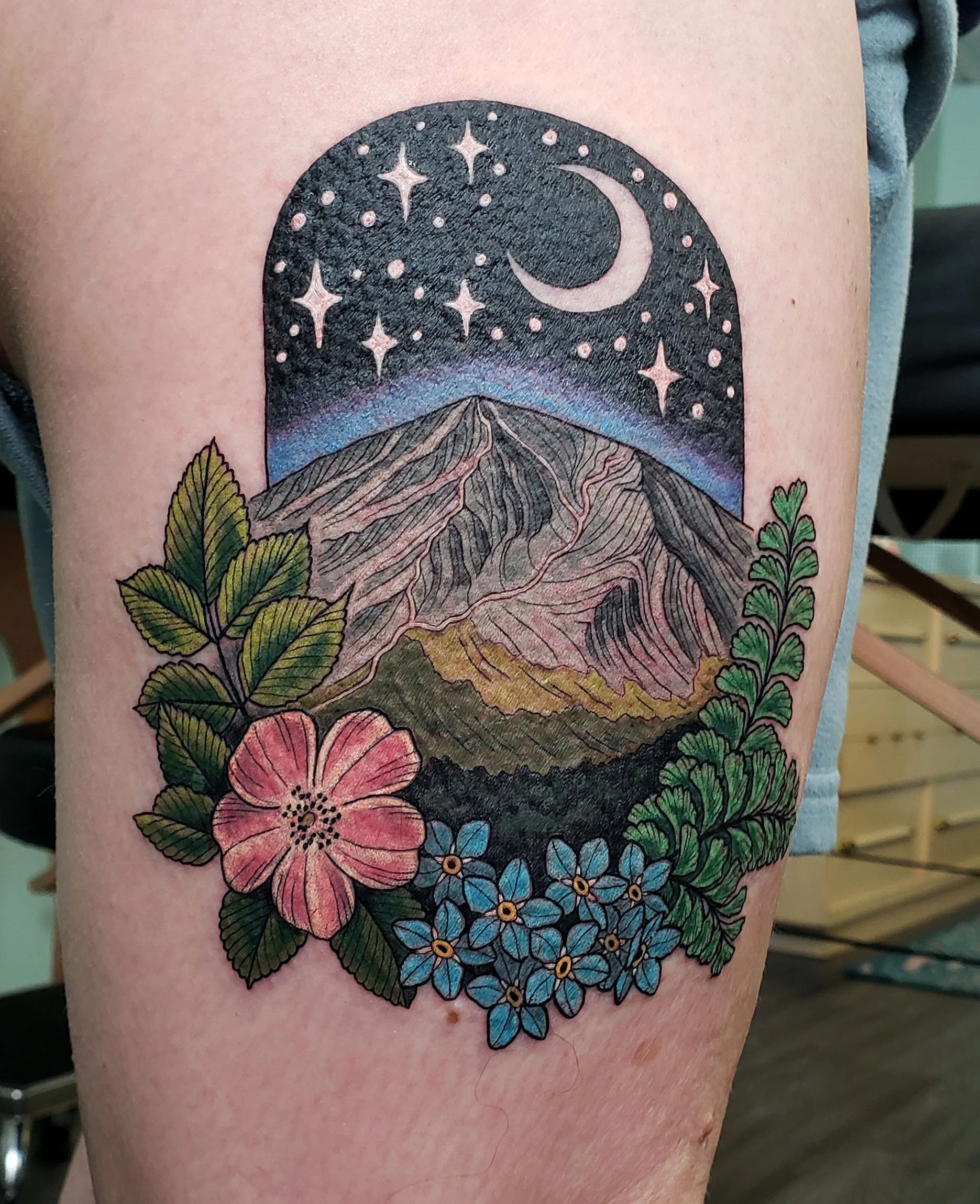 Colorful tattoo of a mountain, moon, stars, and flowers on a person's arm.
