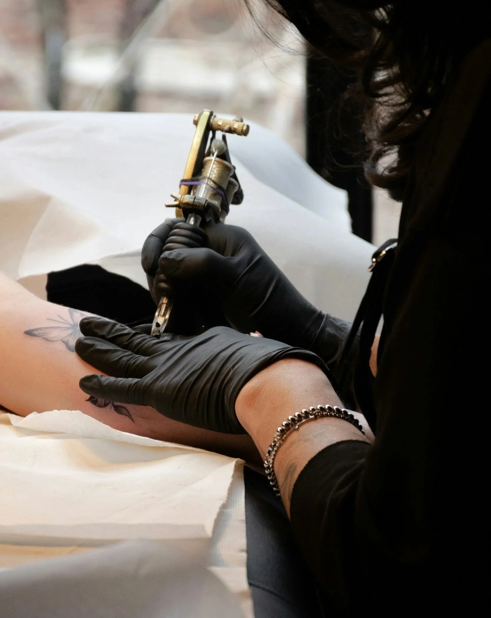 Tattooed arm of person lying on hospital bed, holding tattoo machine with gloved hand, wearing a black bracelet.