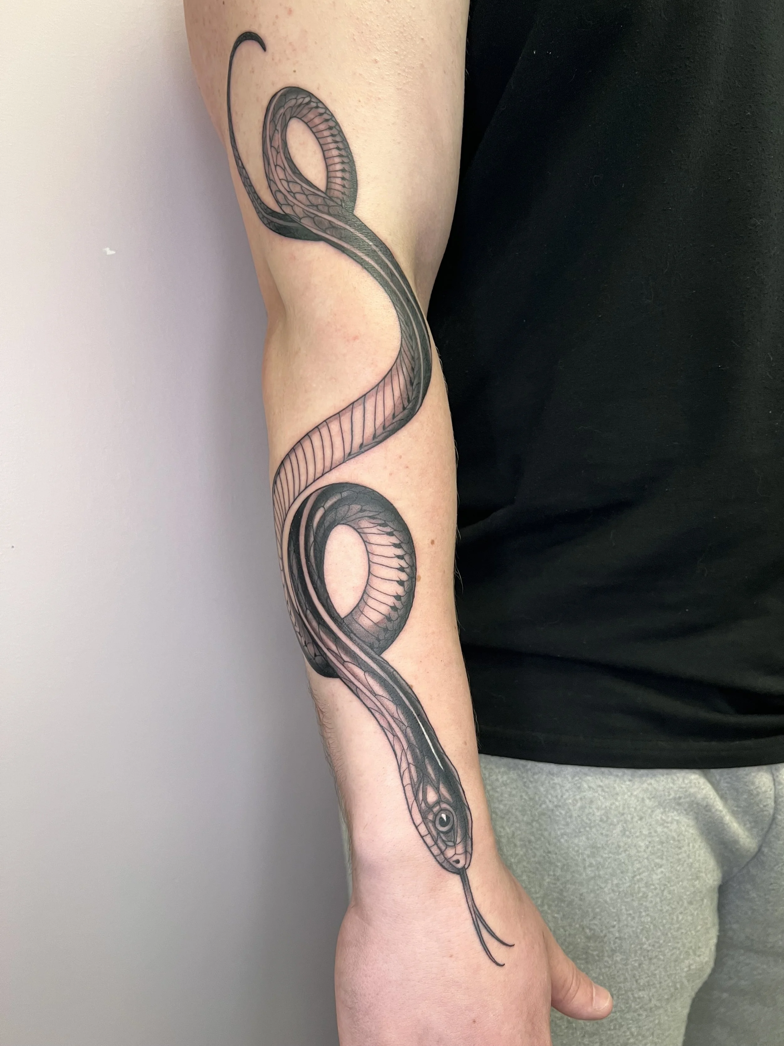 A person's arm with a tattoo of a snake coiled around it, with detailed scales and head near the wrist.