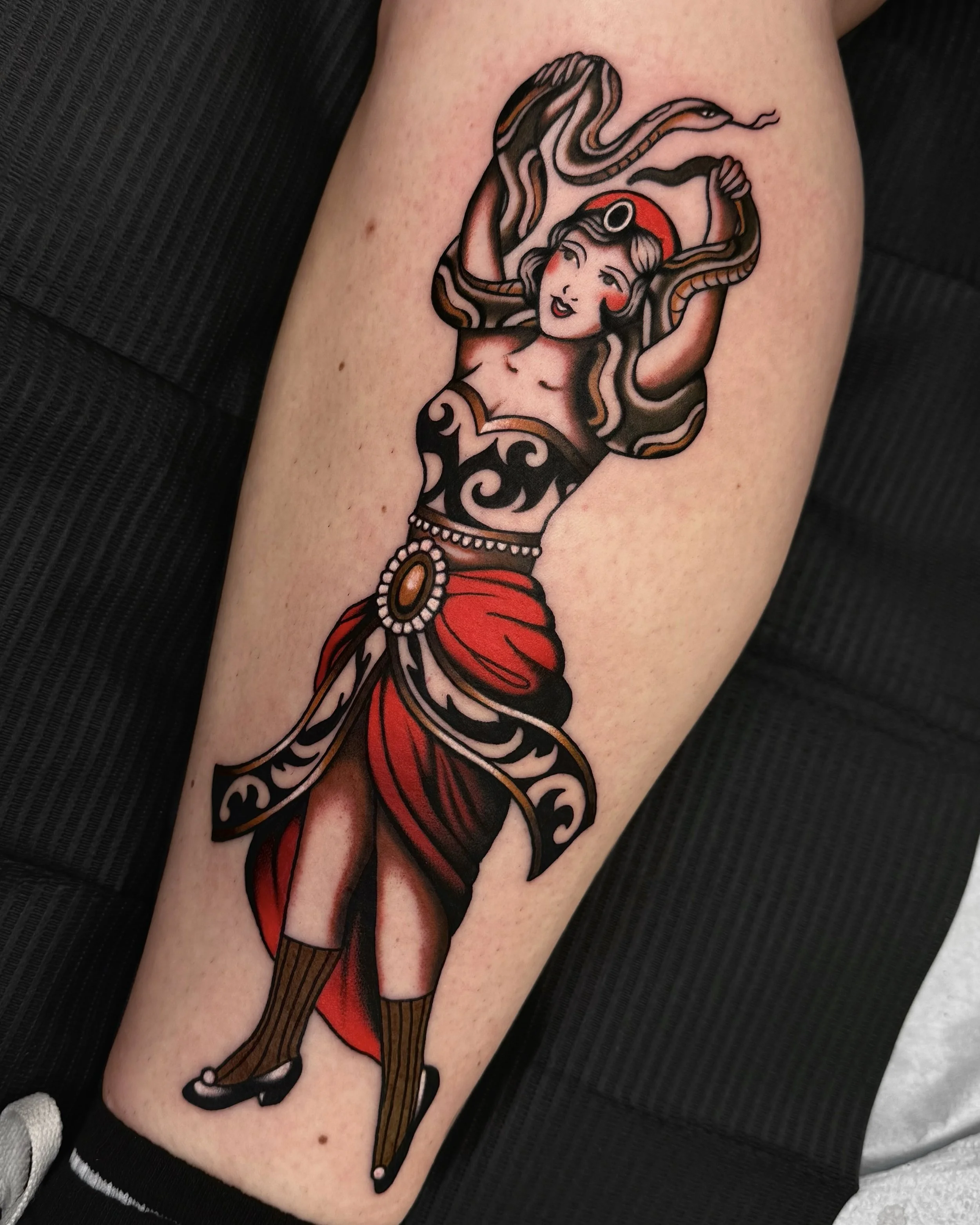 A tattoo of a vintage-style woman with long flowing hair, dressed in a red and black outfit, holding goggles on her head and a scarf, with a snake-like creature entwined around her.