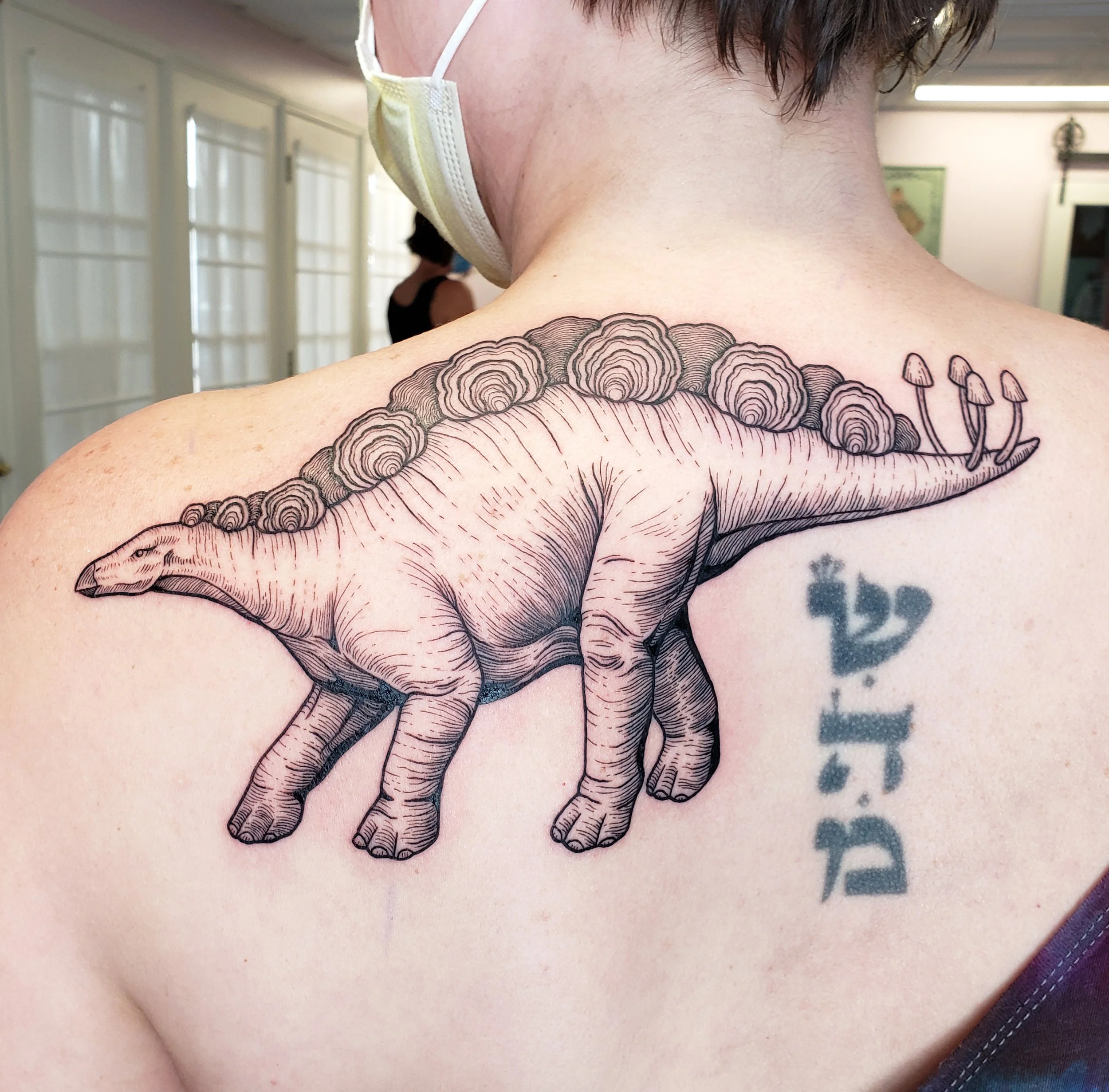Close-up of a person's shoulder with a detailed black ink tattoo of a whale, with part of a face mask visible and a background of a room with windows and a person wearing a mask.