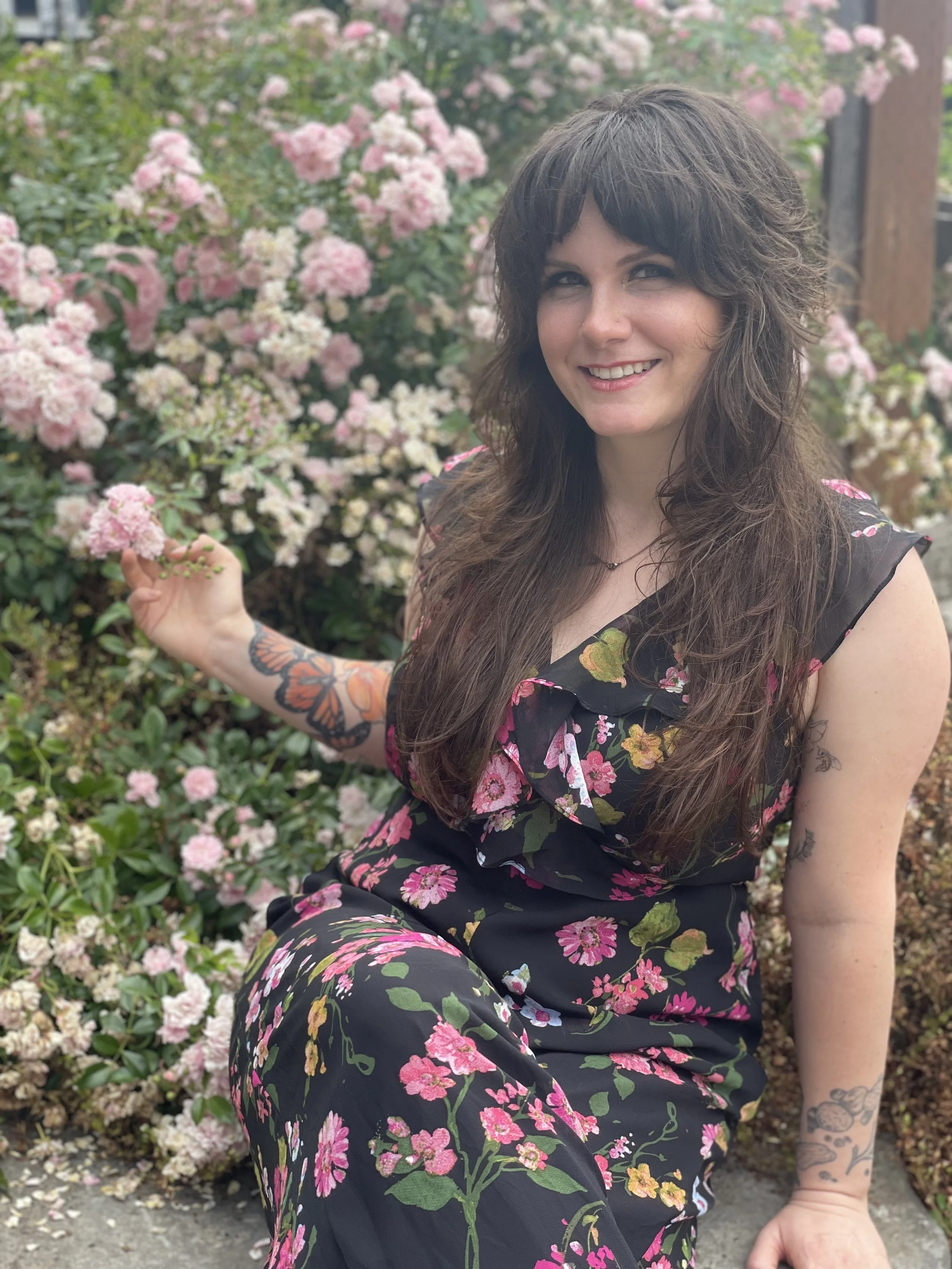 A woman with long brown hair and a floral tattoo on her arm, smiling and sitting outdoors in front of pink and white flowering bushes, wearing a black floral dress.