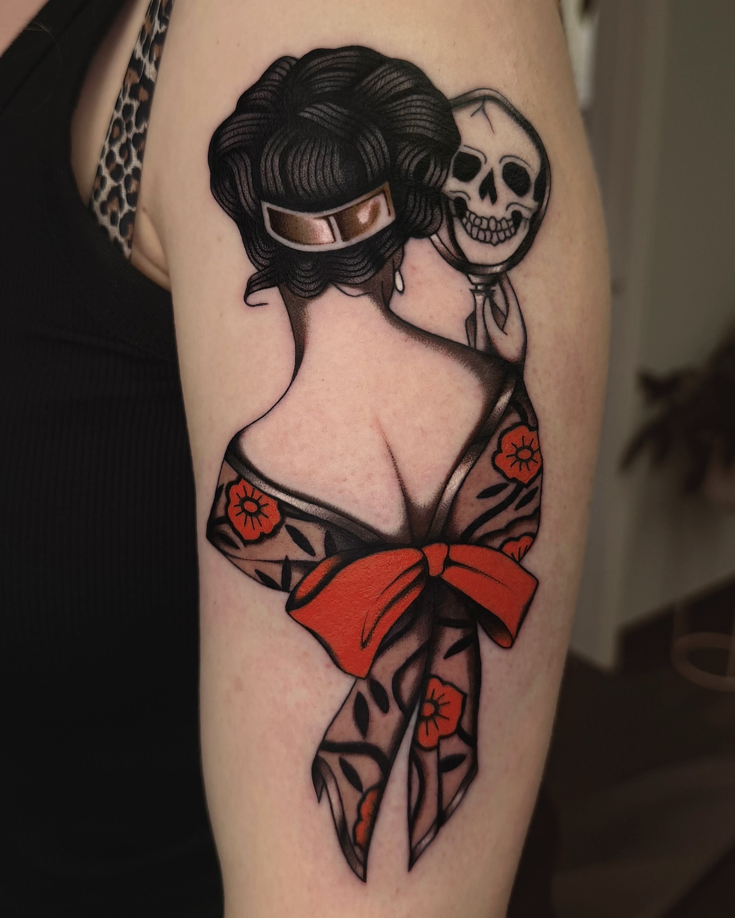 Tattoo of a woman with dark, vintage hairstyle, wearing a floral kimono, holding a skull, with red flowers and a red bow on her kimono.