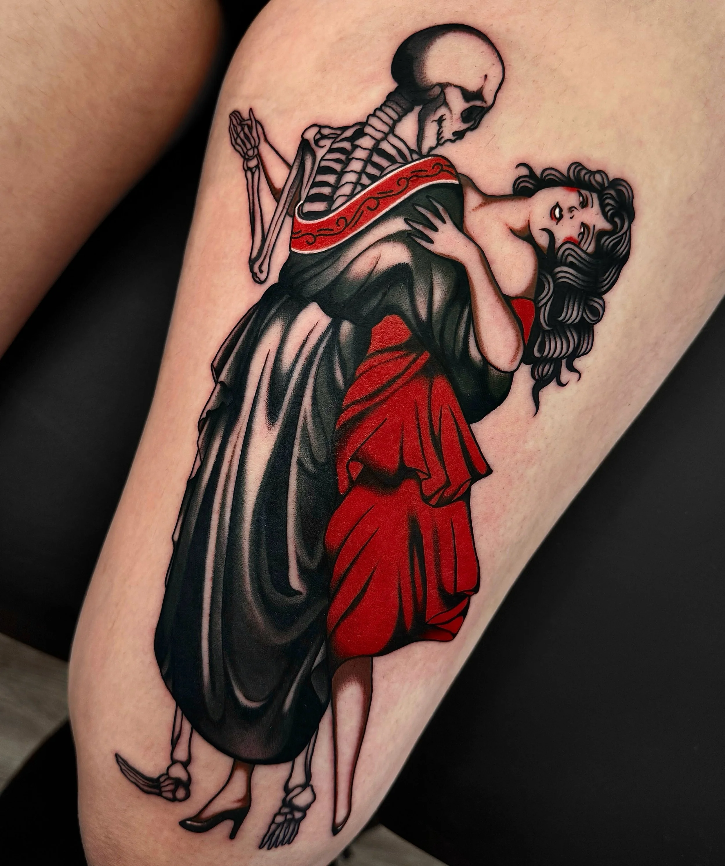 A tattoo of a skeleton and a woman with dark curly hair, red lipstick, and a red dress. The skeleton appears to be holding a bow and arrow, with the arrow pointing toward the woman.