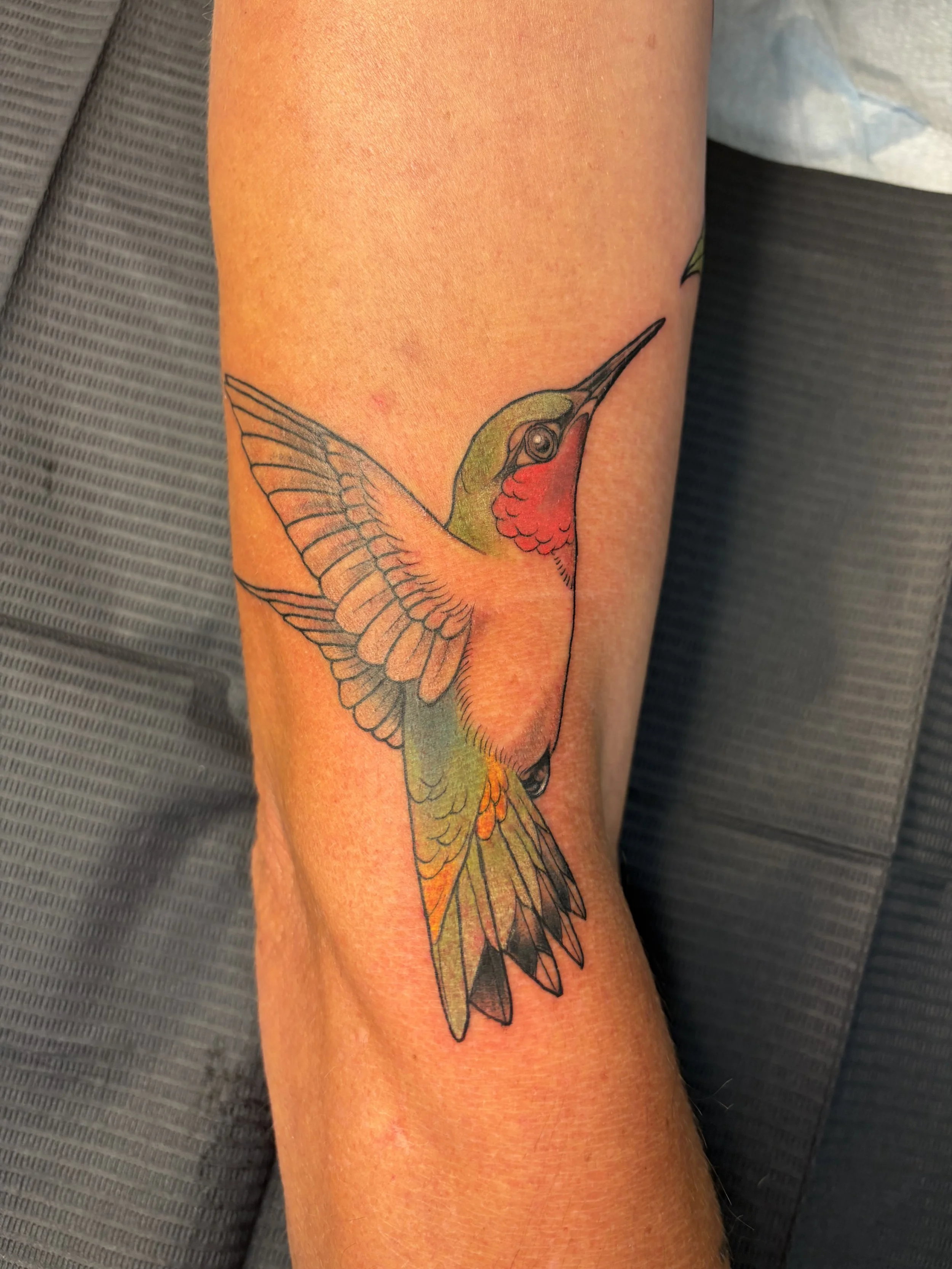 Colorful tattoo of a hummingbird with red throat patch and green head on a person's arm.