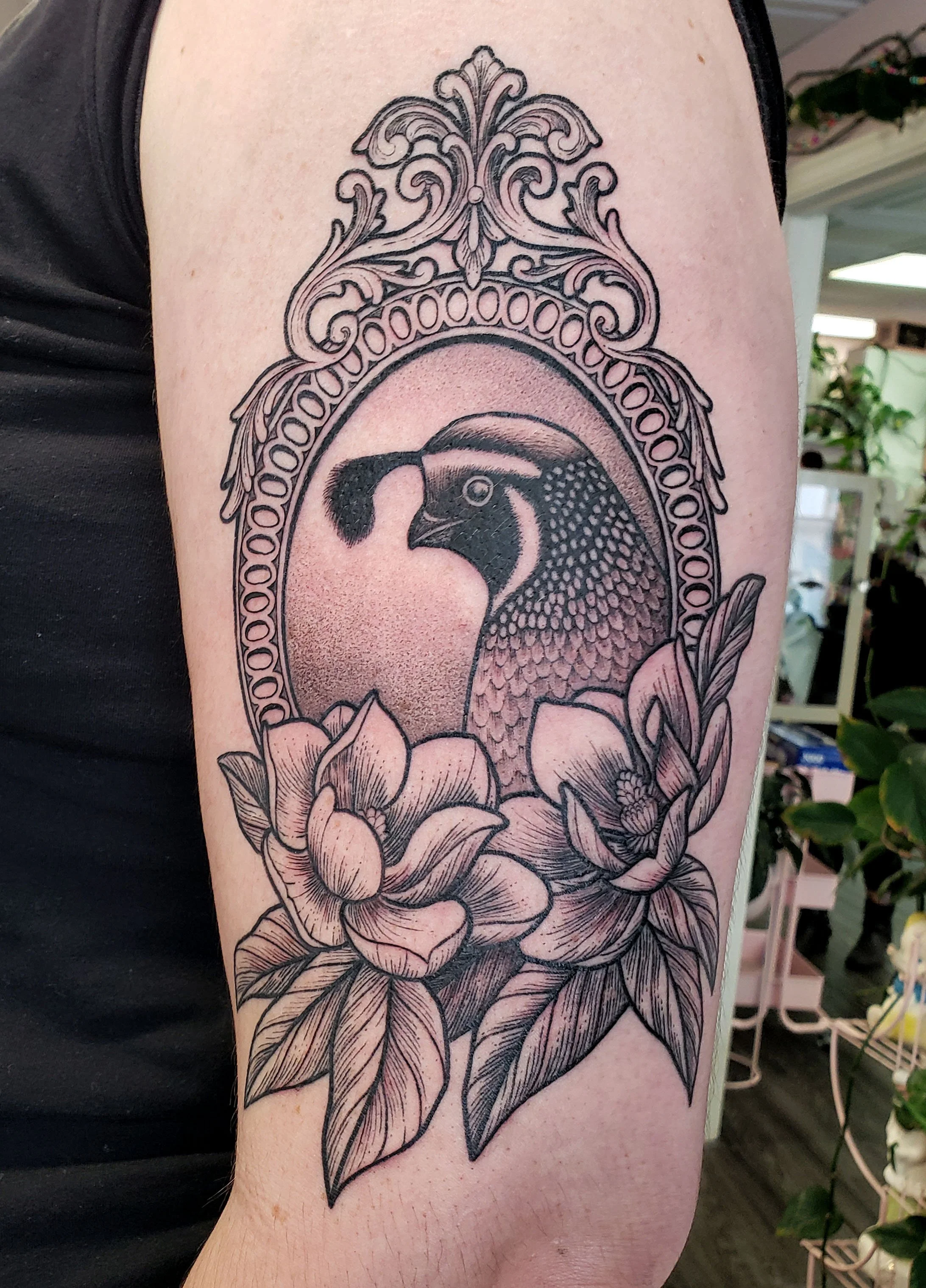 Black and gray tattoo of a quail framed in an ornate oval with floral details on the arm.
