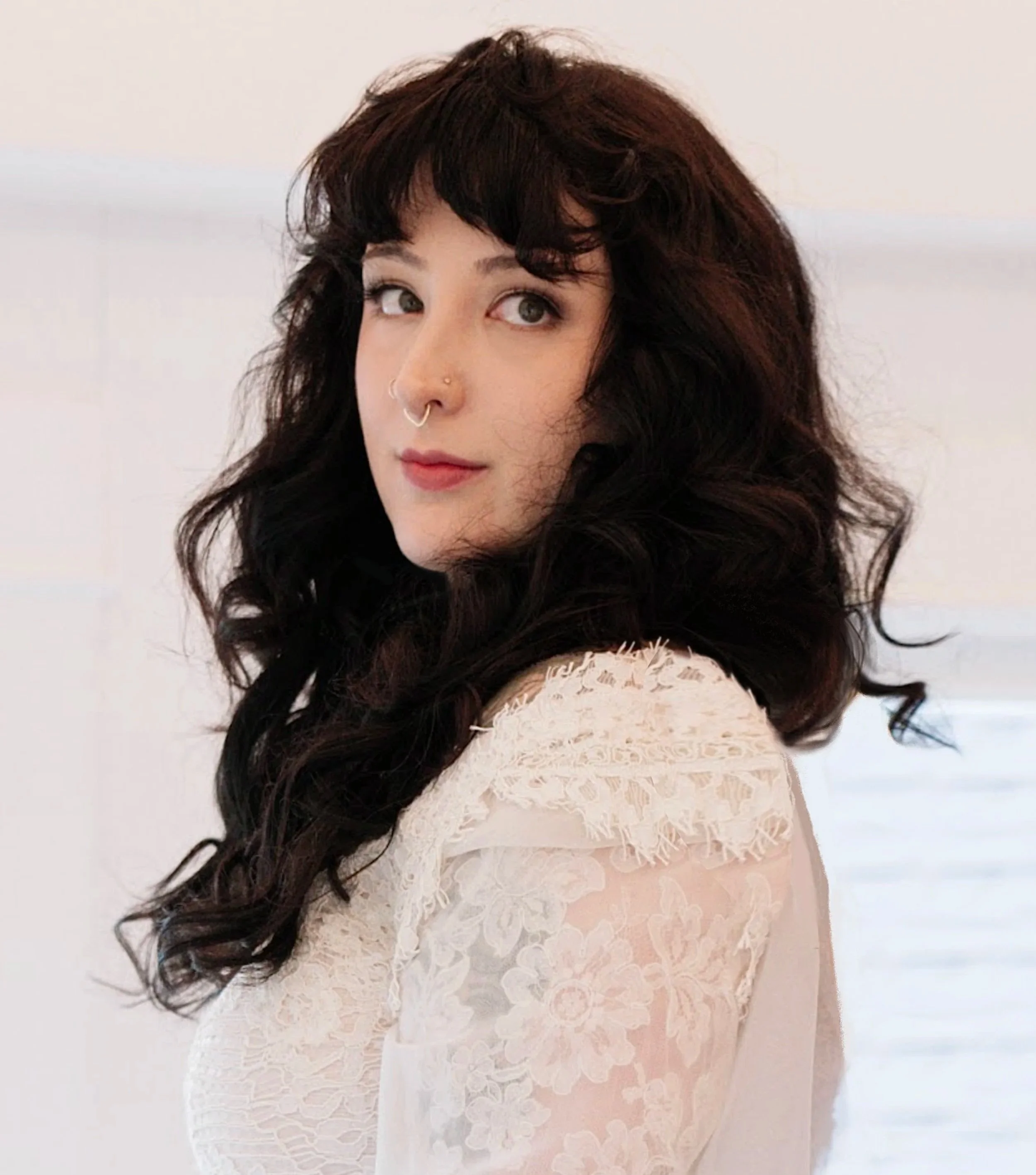 A woman with long dark wavy hair wears a light-colored lace dress, standing against a simple light background.