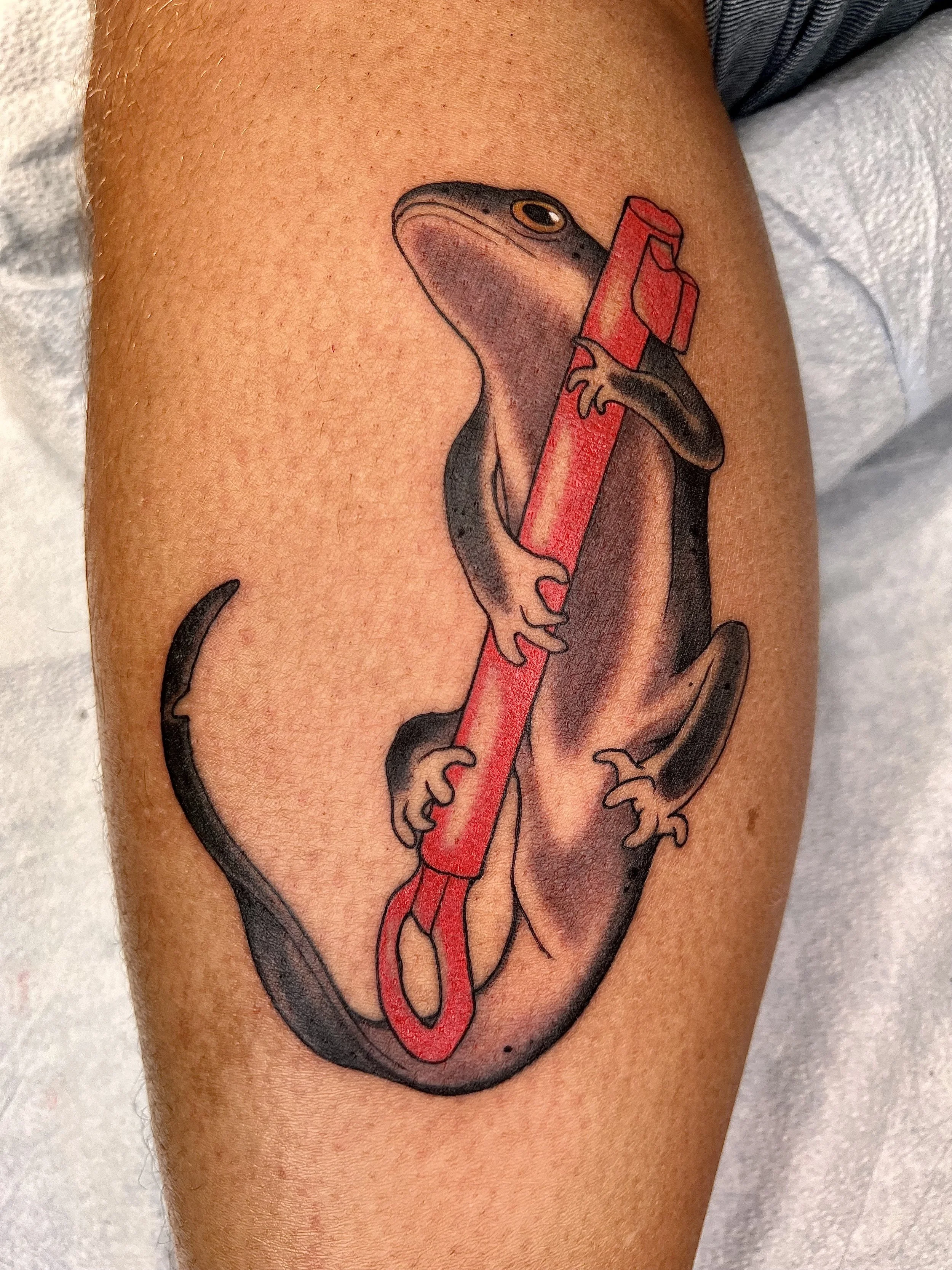 A tattoo of a cartoon-style lizard holding a red pen, with the lizard's tail forming an incomplete circle around it.