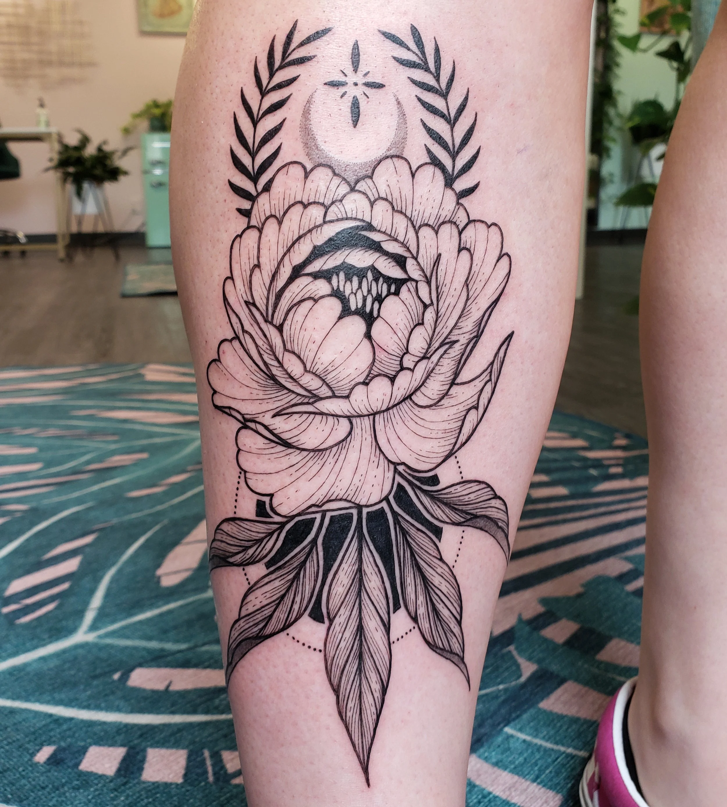 Black outline floral tattoo on person's thigh featuring a large flower, leaves, and a crescent moon with a star and decorative dots above.