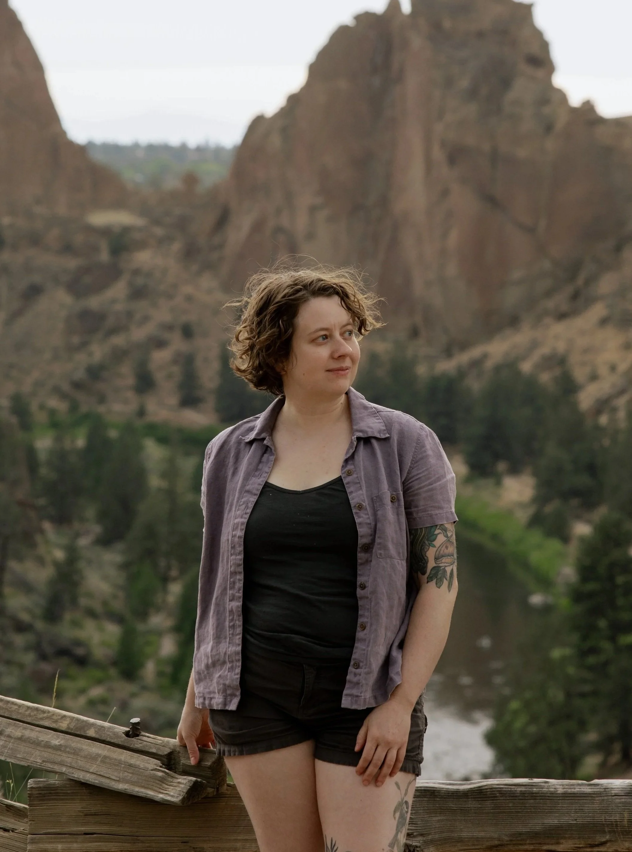 A woman with short curly hair wearing a black top and purple shirt standing outdoors near a wooden fence at Smith Rock, with a lake and large rocky cliffs in the background.