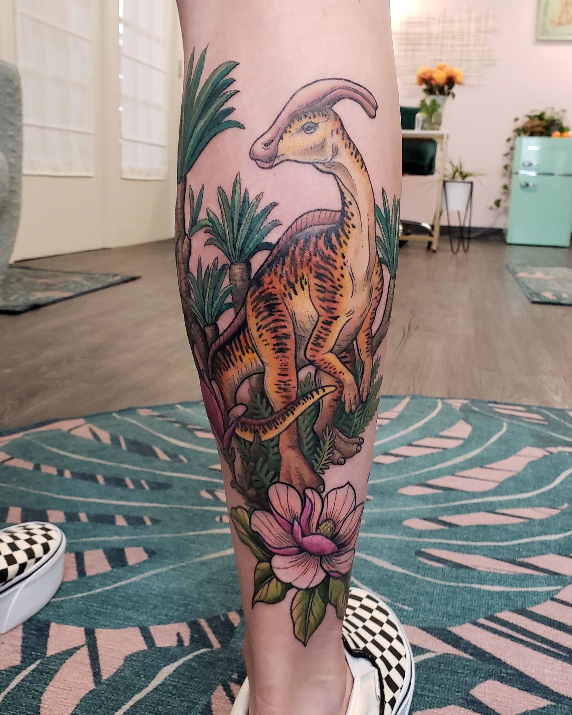 A colorful tattoo of a dinosaur, surrounded by tropical plants and flowers, on a person's lower leg.