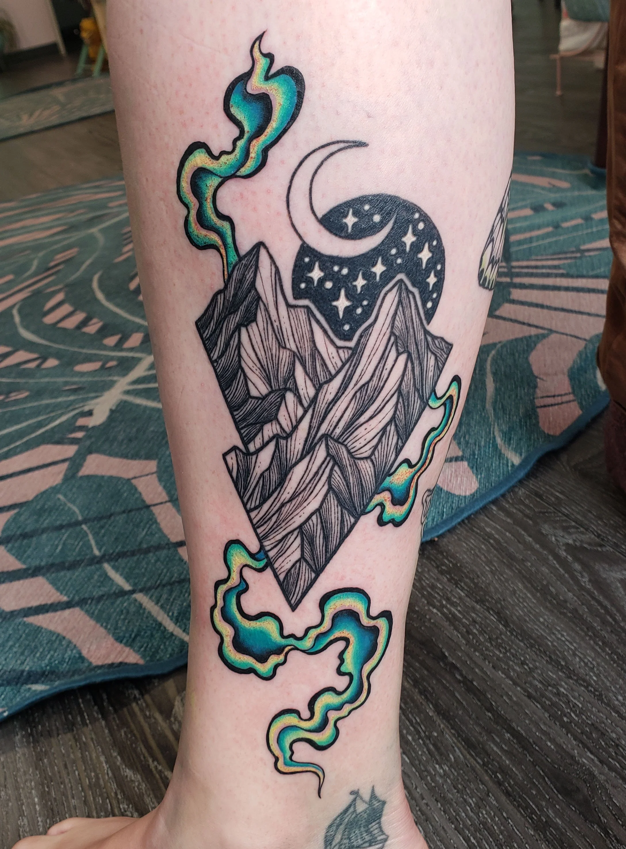 Tattoo of mountains with a starry night sky, moon, and colorful smoke or cloud above on person's leg.