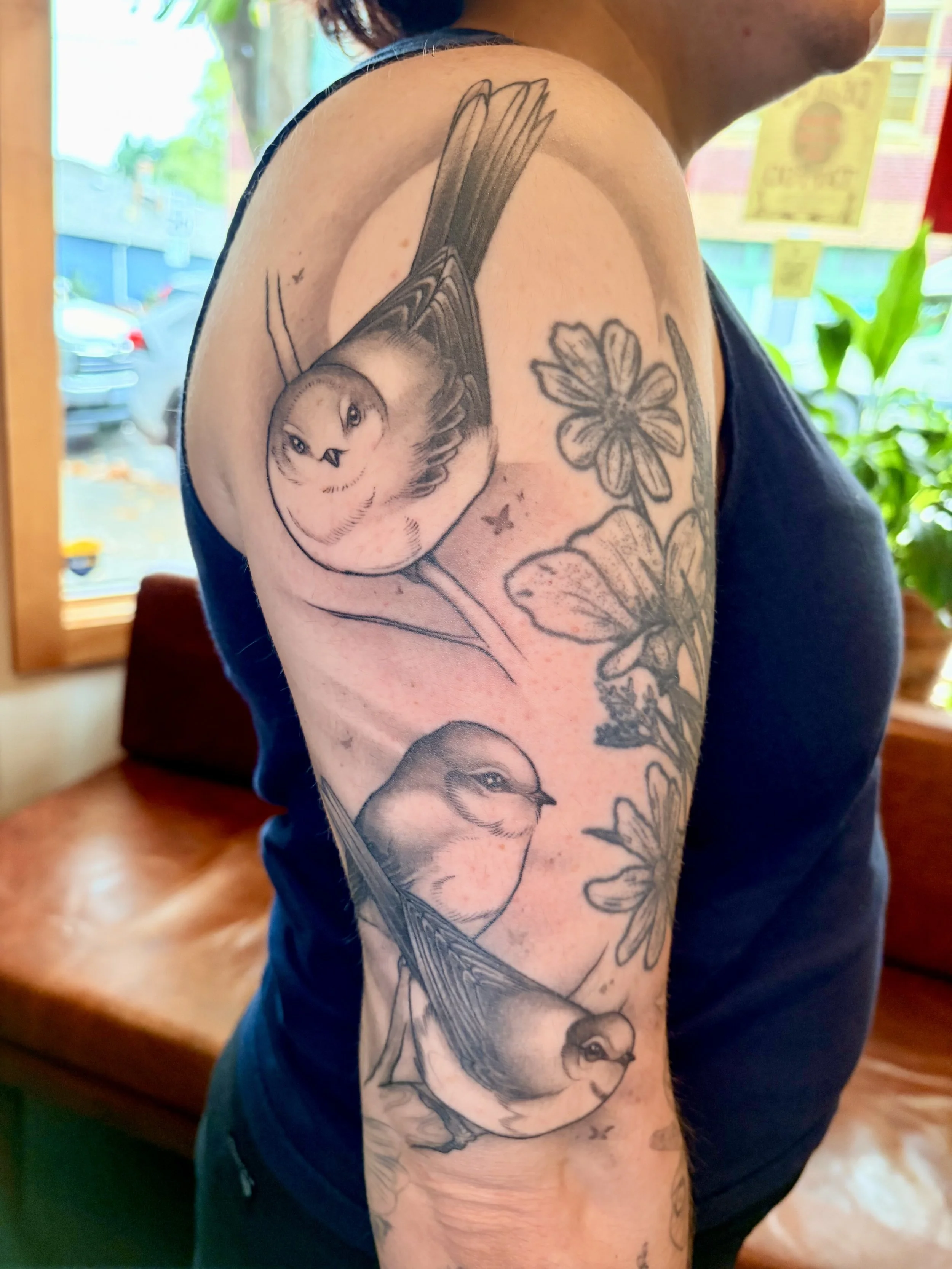 Close-up of a person's upper arm and shoulder with a detailed black and gray tattoo of a bird and flowers, in a bright indoor setting.