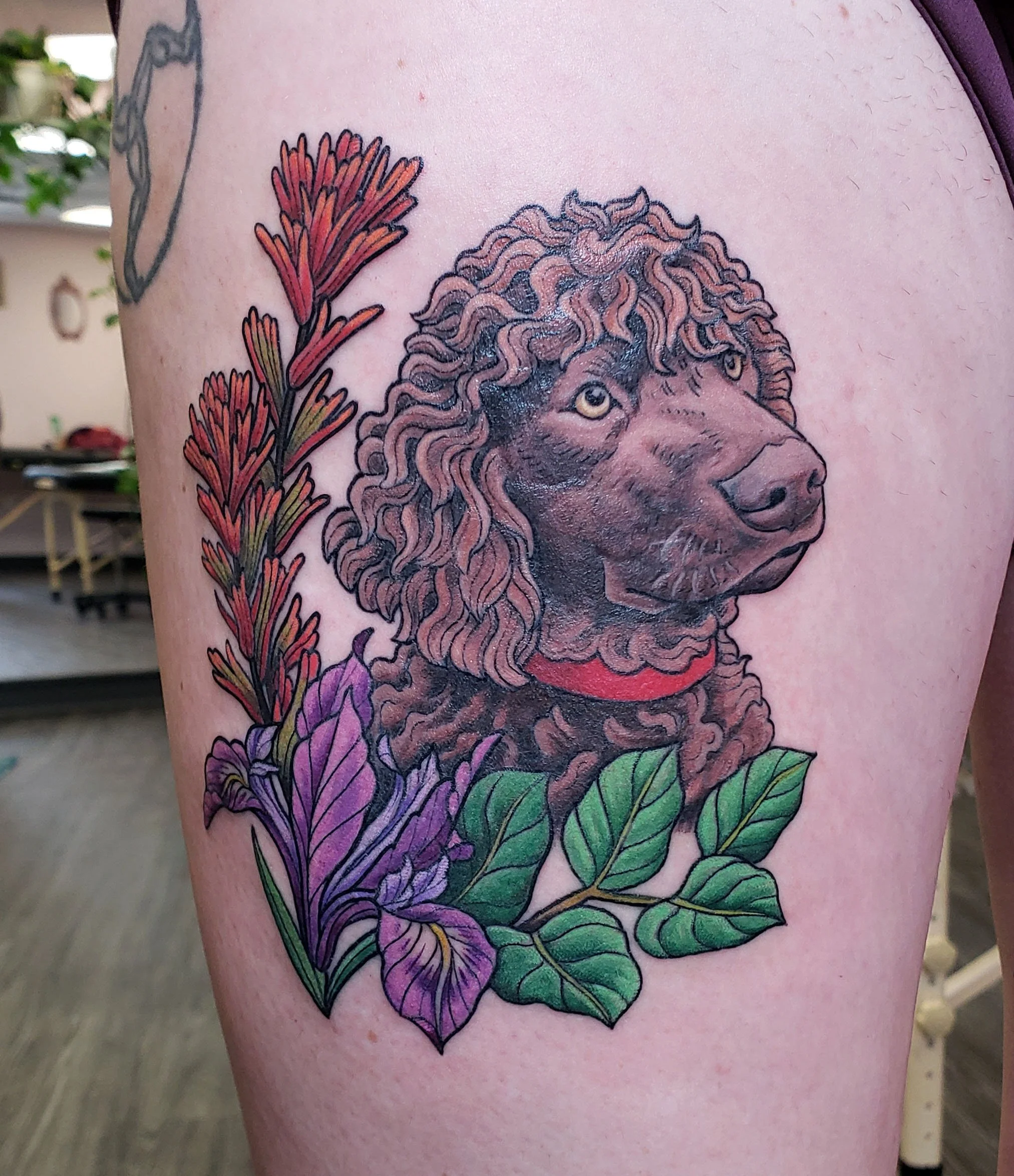 Colorful tattoo of a poodle dog's face surrounded by large purple flowers and green leaves on person's upper arm.