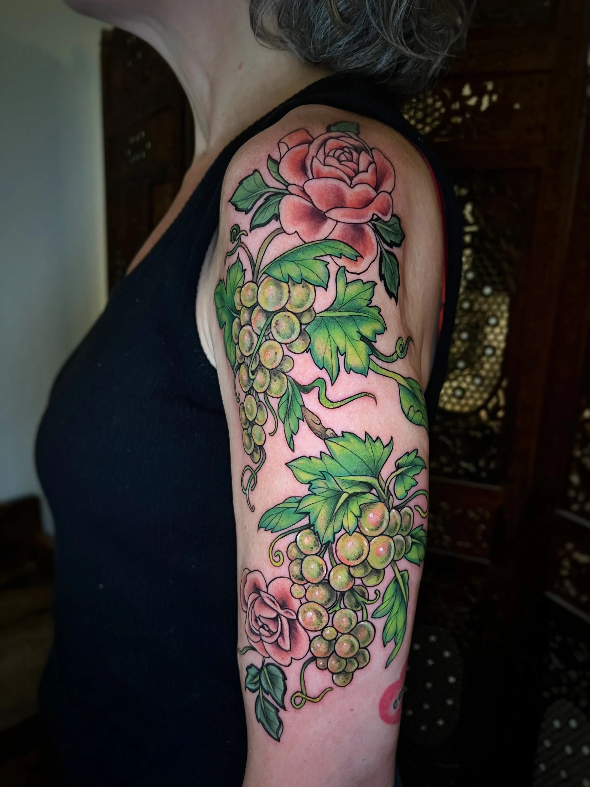 Colorful tattoo of a pink rose, green leaves, and a cluster of grapes on a woman's upper arm.