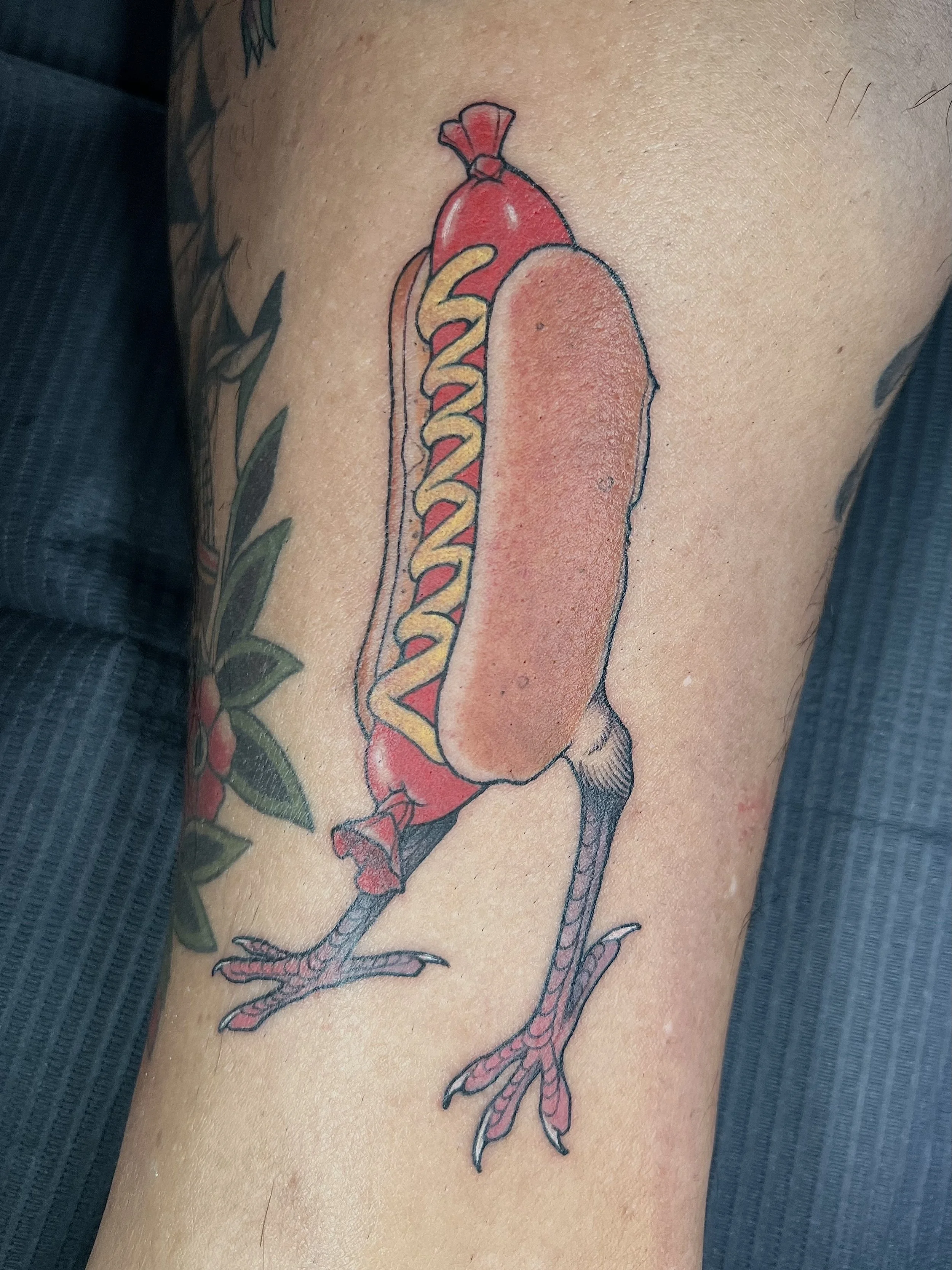 Tattoo of a chicken with a hot dog bun as its body, a sausage with mustard and ketchup as its neck, standing on two legs, with leaves in the background.