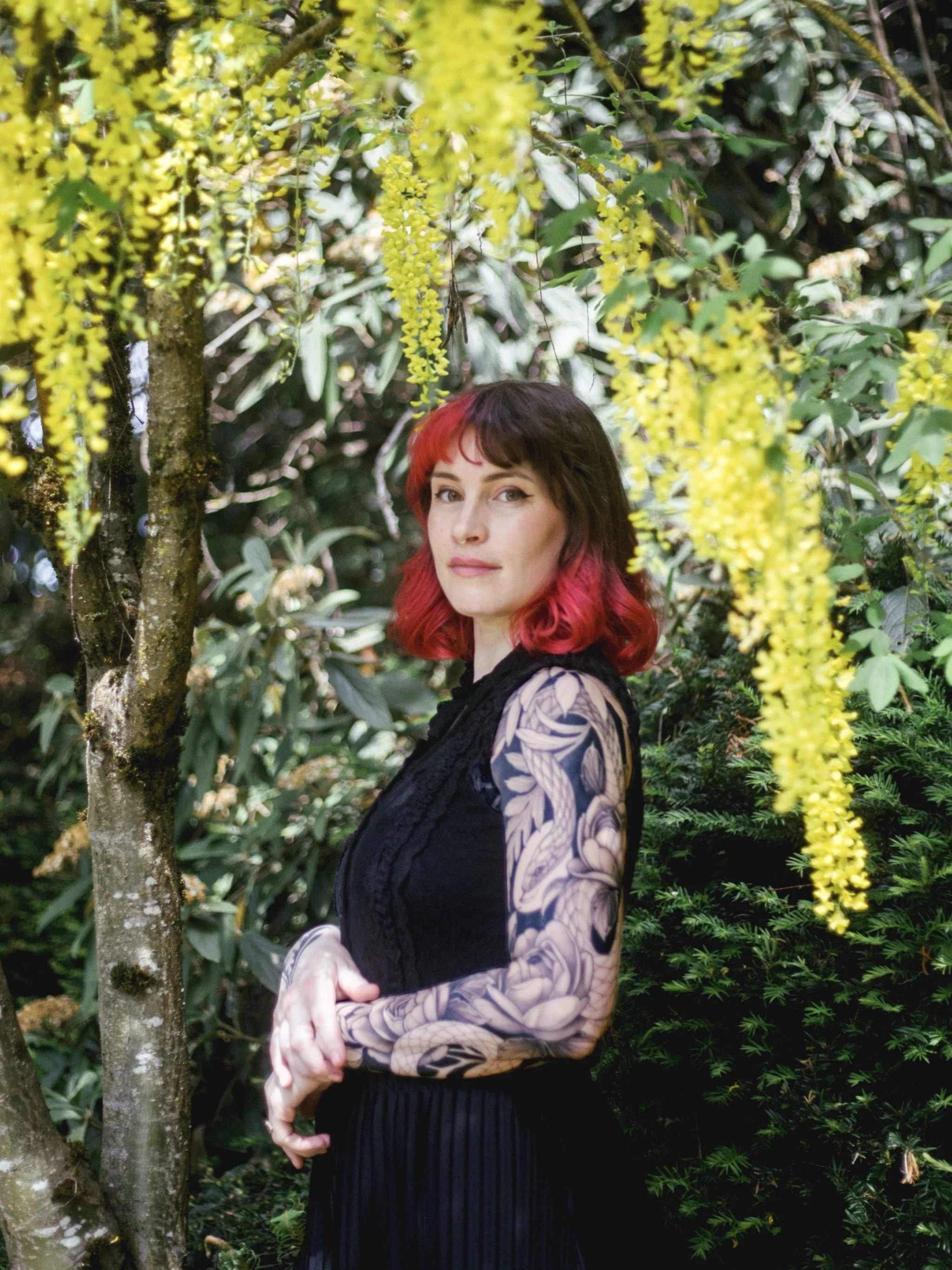 A woman with red and brown hair standing among green foliage and yellow hanging flowers, wearing a black lace dress with floral and geometric patterned tattoos down her arm