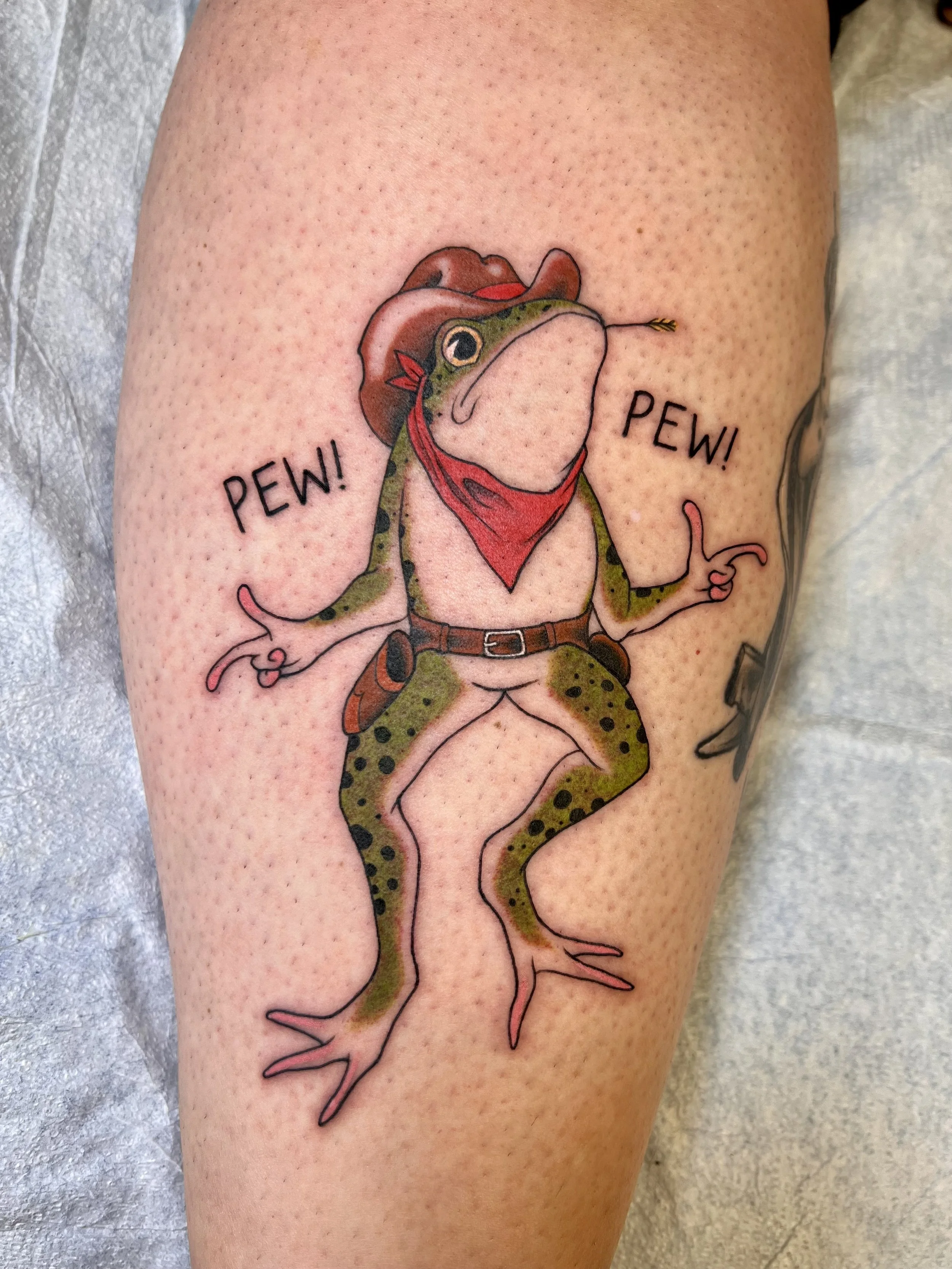 Tattoo of a frog wearing a cowboy hat and red bandana, with the words "PEW! PEW!" on either side.