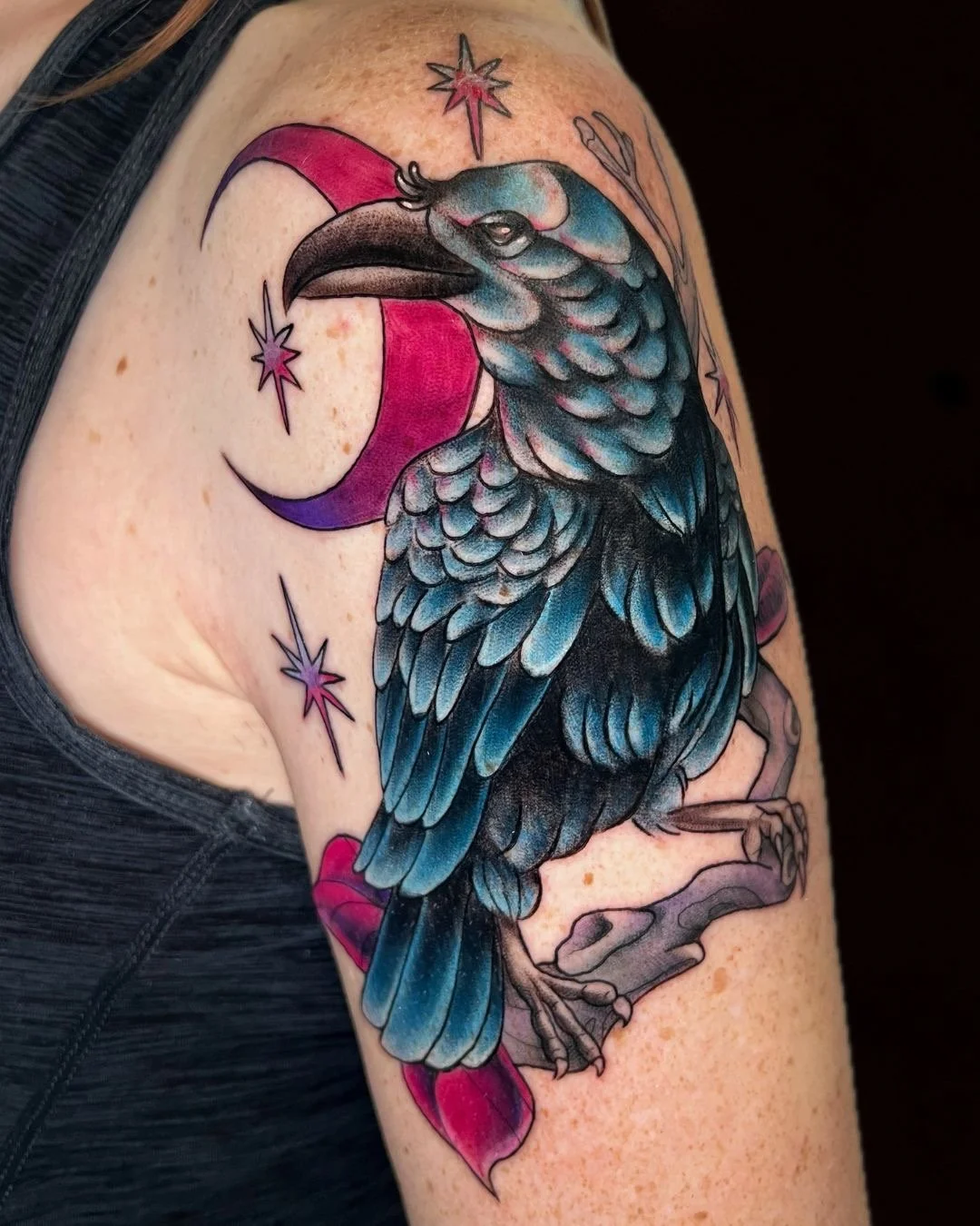 Colorful tattoo of a blue and black bird, possibly a crow or raven, perched on a branch. The background features a red and purple crescent moon and three purple star-shaped designs.