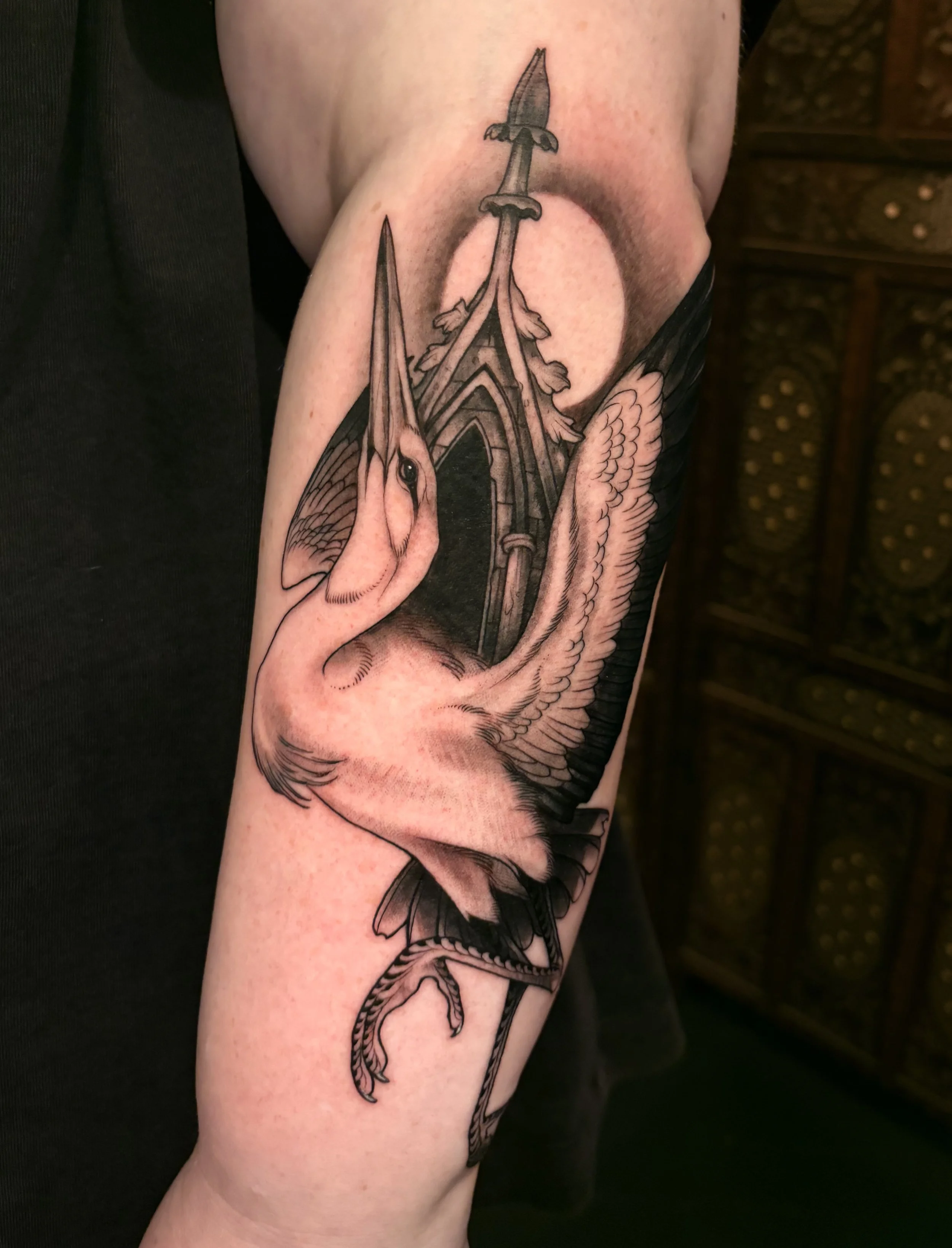 Illustrative black and gray tattoo of a crane with a cathedral window in the background. The tattoo is done with bold high-contrast shading on an arm