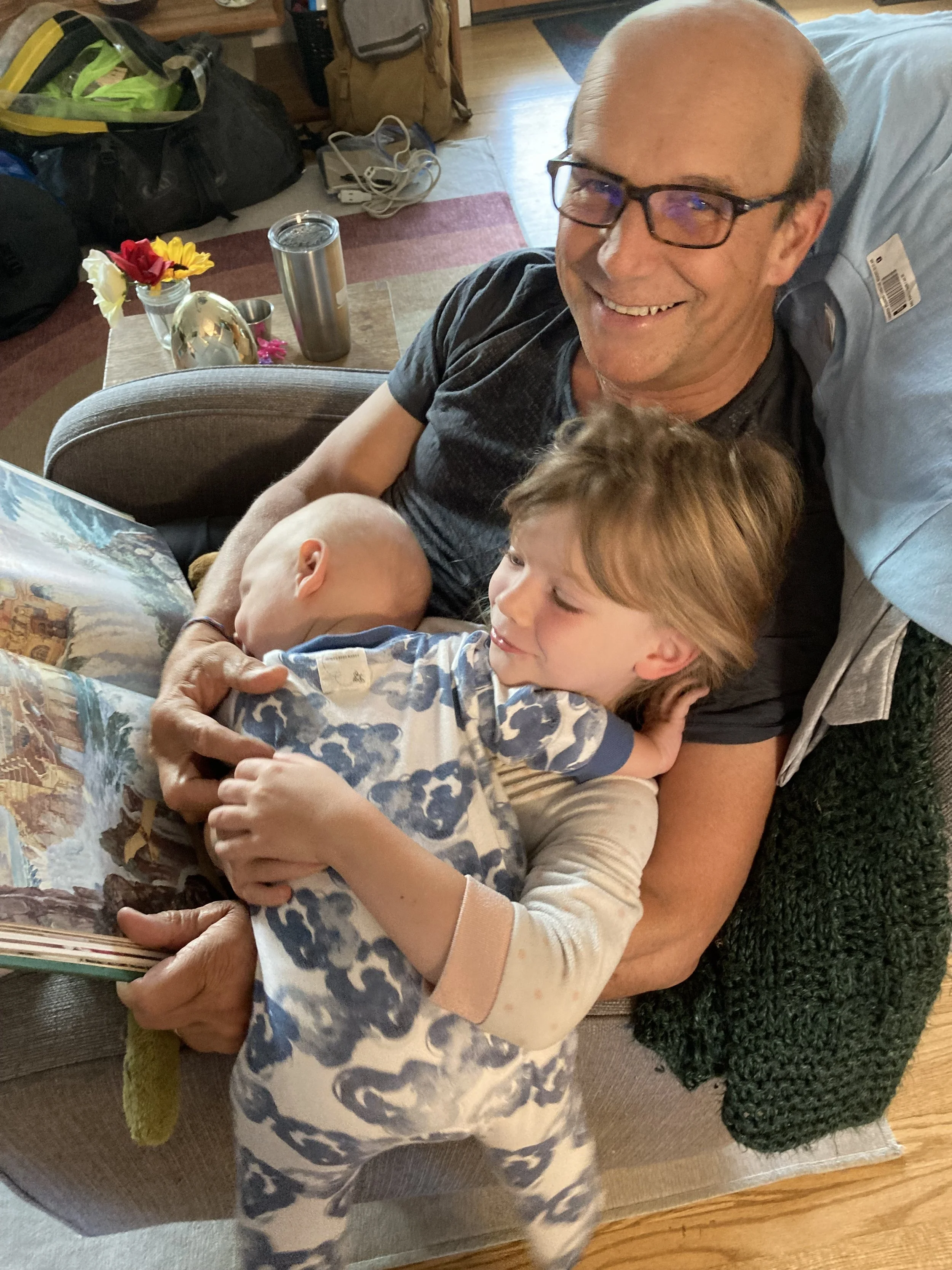 A man with glasses smiling on a couch, holding two children who are resting on his lap, one with their head on his chest and the other with their head on his shoulder, in a cozy living room.