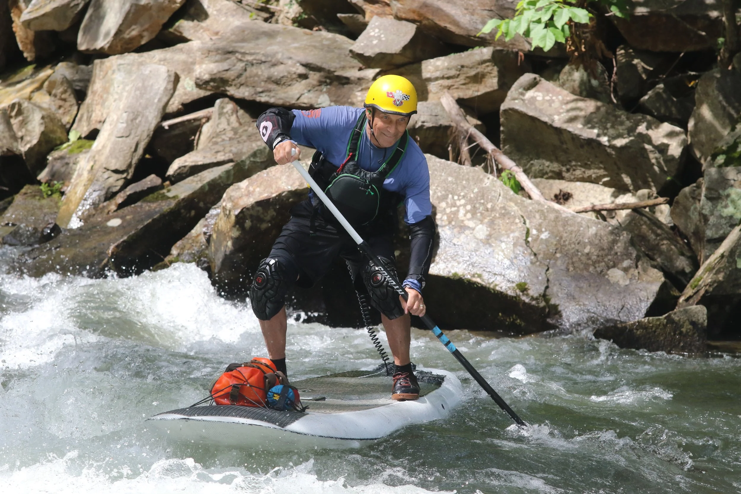 A man in a blue shirt and black shorts wearing a yellow helmet paddleboarding in a river surrounded by rocks and trees.