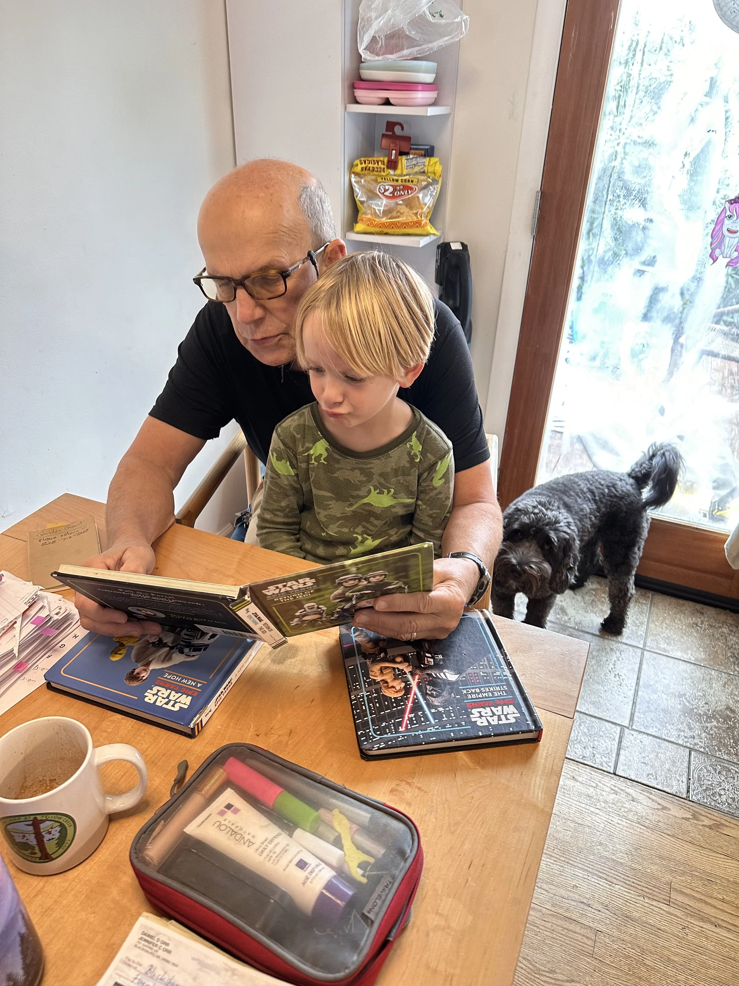 A man with glasses and a young boy sit at a wooden table, reading magazines. On the table, there are two coffee mugs, a red and gray pencil case, and another magazine. In the background, a black dog stands near a glass door, and there is a shelf with dishes and snacks behind them.