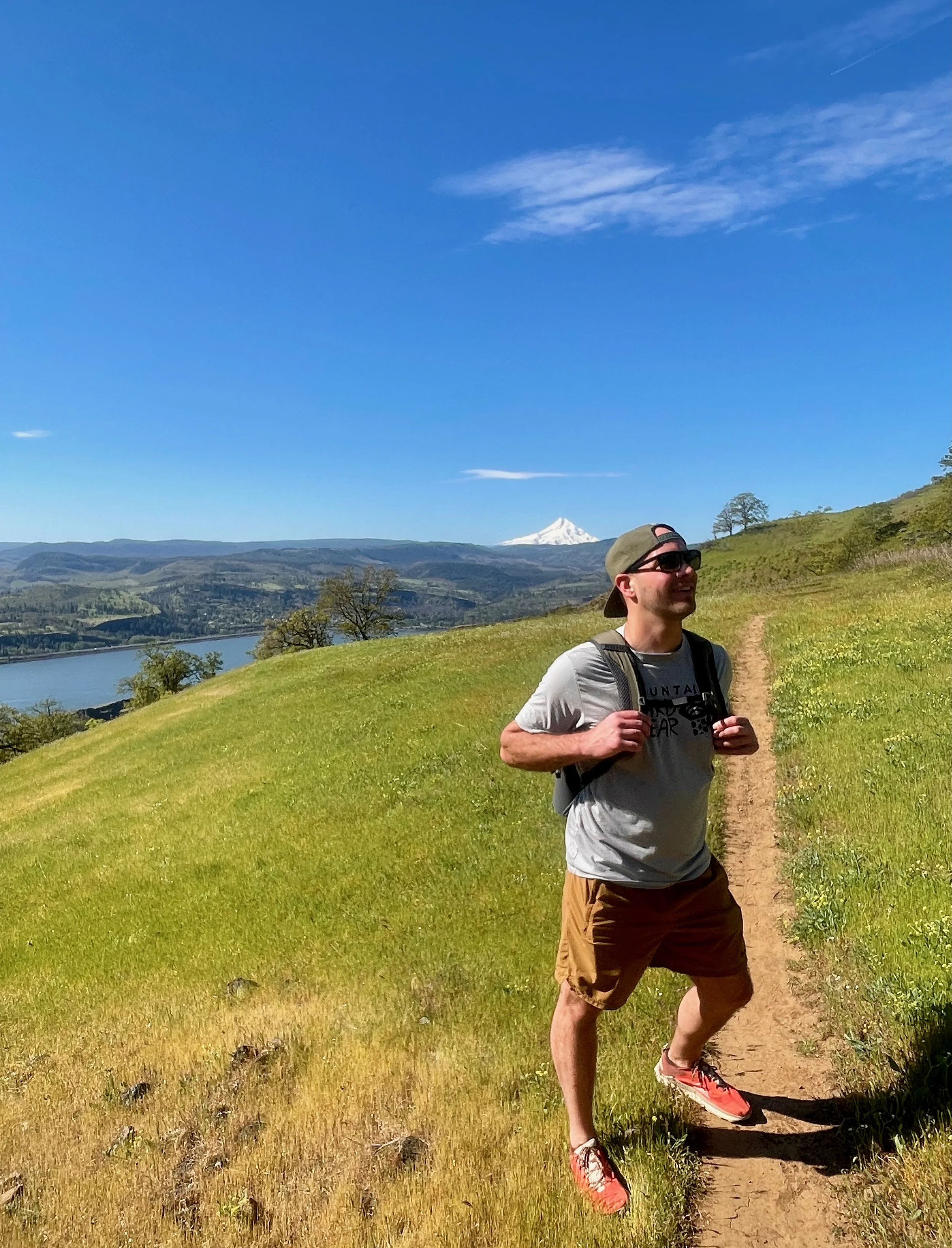 A man wearing sunglasses, a baseball cap, a backpack, a gray t-shirt, and brown shorts, hiking or jogging on a dirt trail through a grassy landscape with hills and a mountain in the background, under a blue sky with a few clouds.