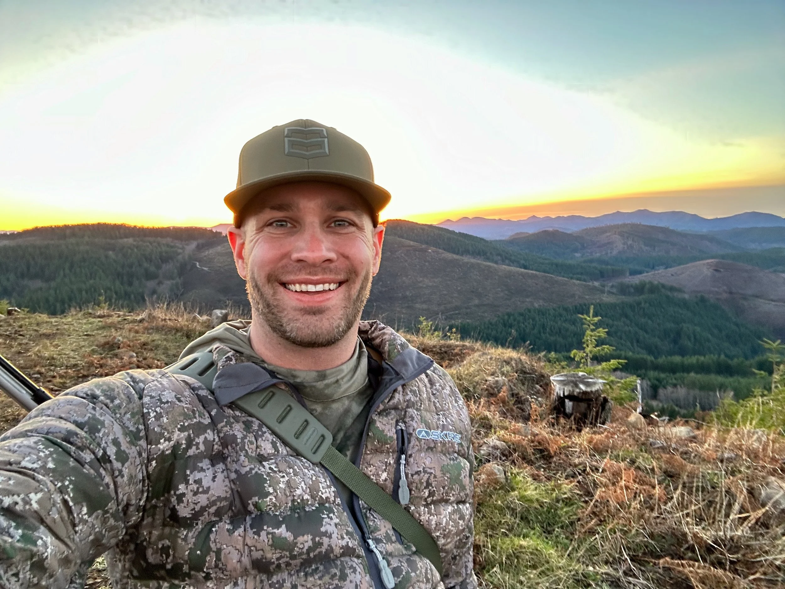 A man taking a selfie outdoors during sunset, wearing camouflage gear and a tan cap, with mountains and a sunset in the background.