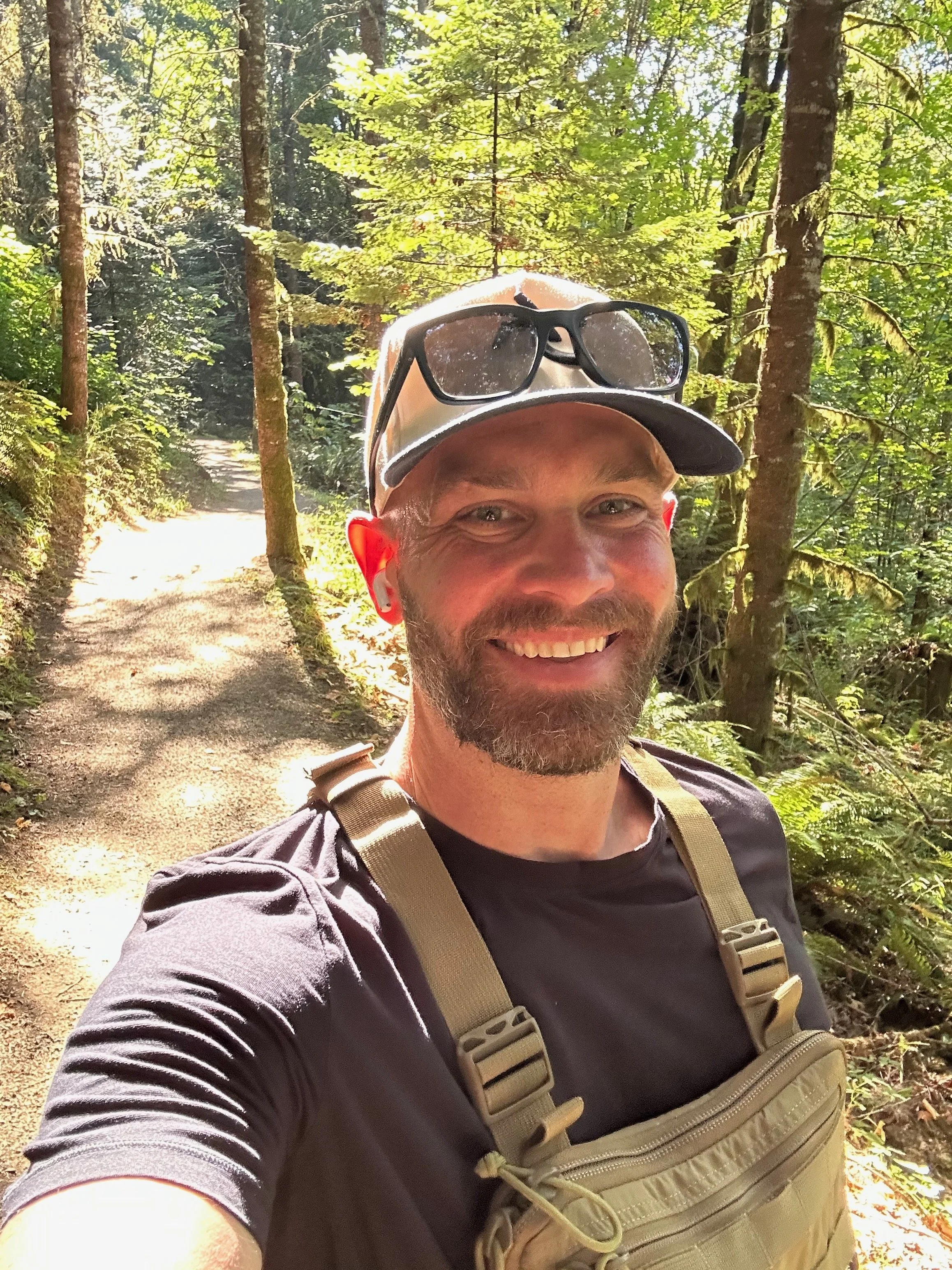 A man smiling and taking a selfie in a lush green forest with tall trees and a dirt trail.