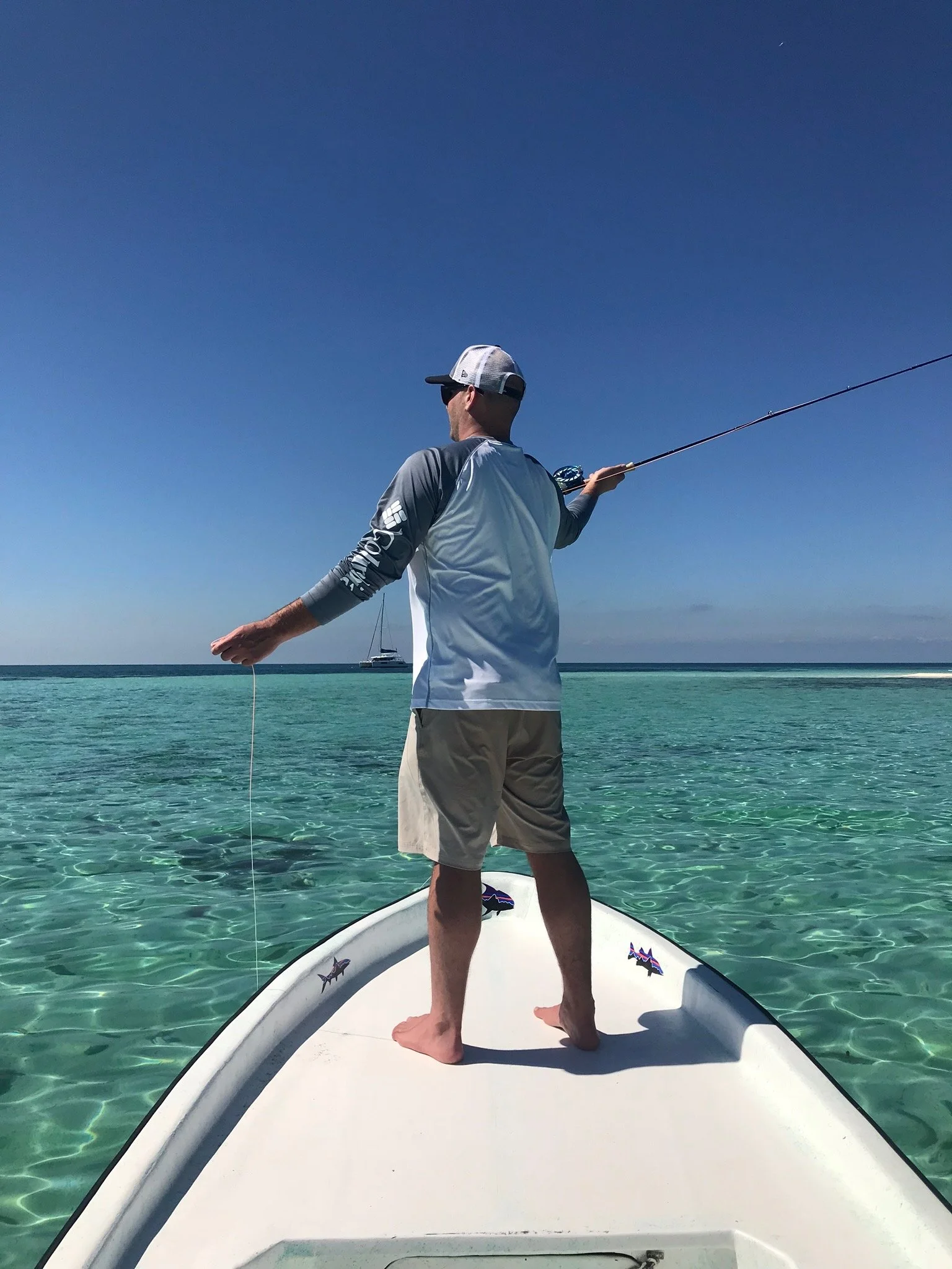 Man standing on a boat with a fishing rod, overlooking the clear ocean with a boat in the distance under a blue sky.
