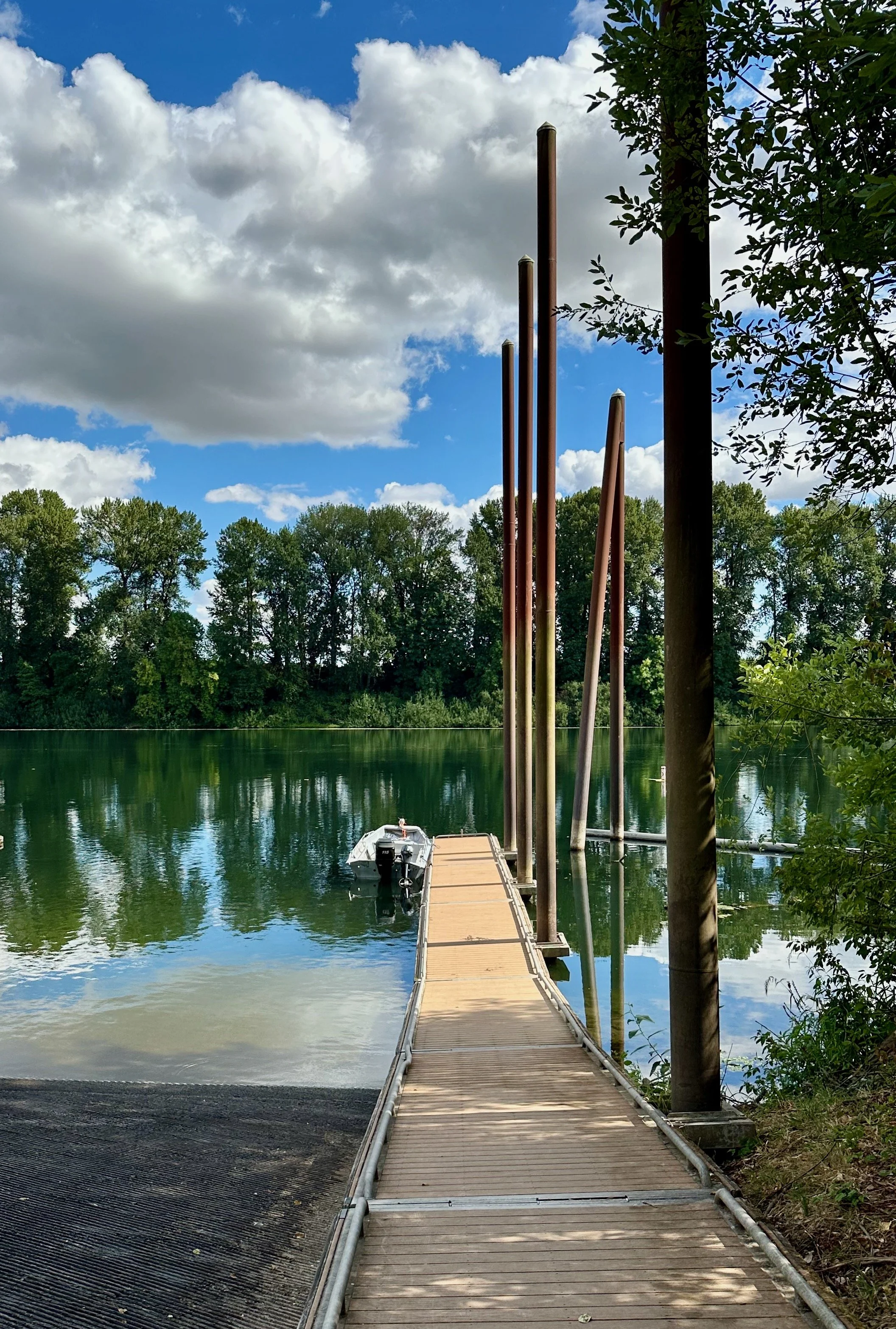 A wooden dock extending into a calm lake, with a boat tied at the end, green trees in the background, and a partly cloudy sky overhead. There are tall poles along the dock and trees on the right side.