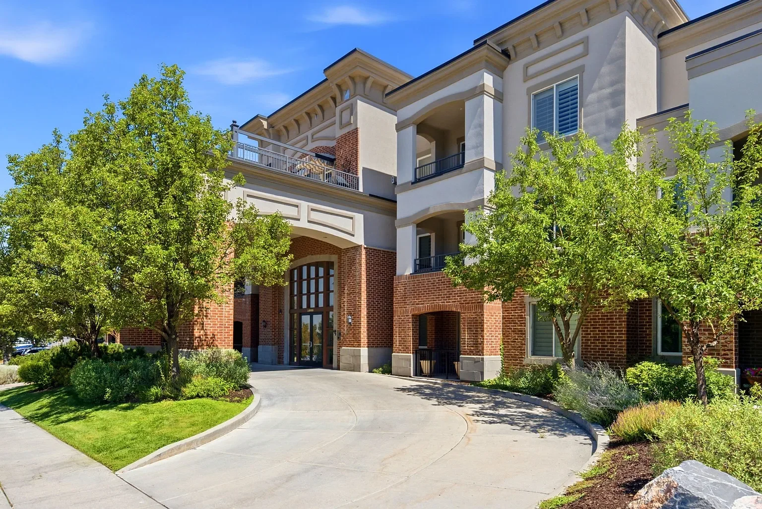 Exterior view of a modern apartment building with brick and stucco architecture, surrounded by green trees and landscaped bushes, under a clear blue sky.