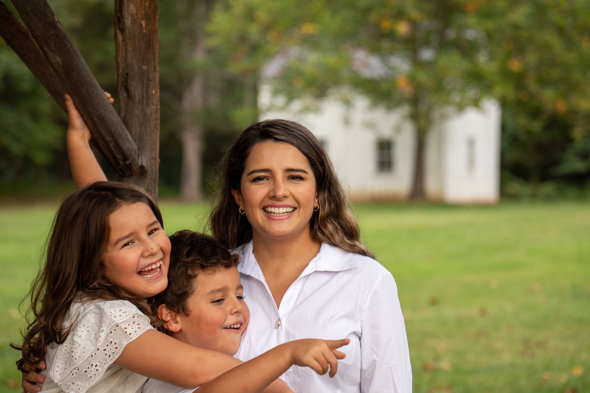 Mother laughing with her son and daughter, the girl pointing at something off camera, white farmhouse behind them.