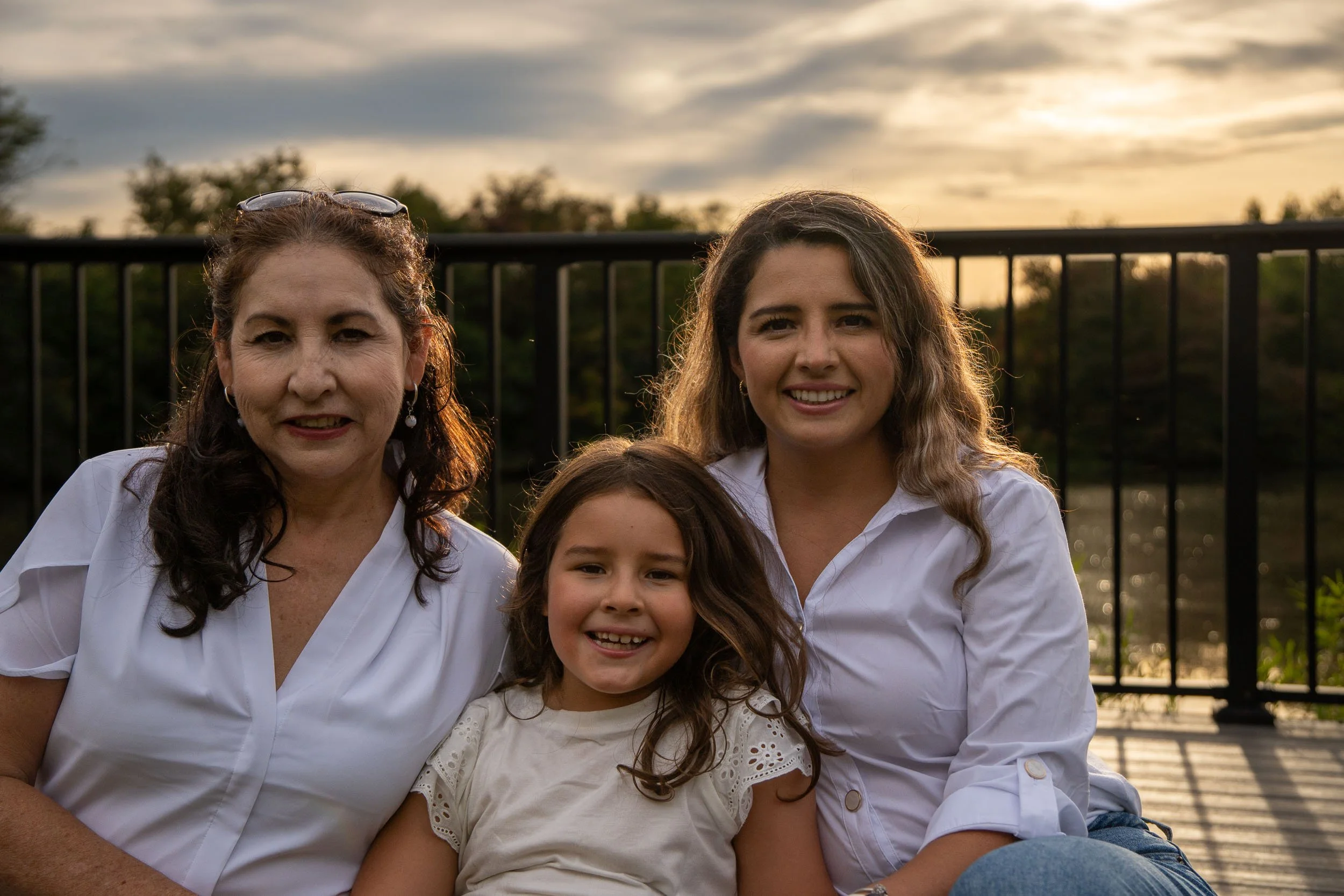 Grandmother, mother and young daughter sitting together and smiling at a lakeside overlook in warm evening light.