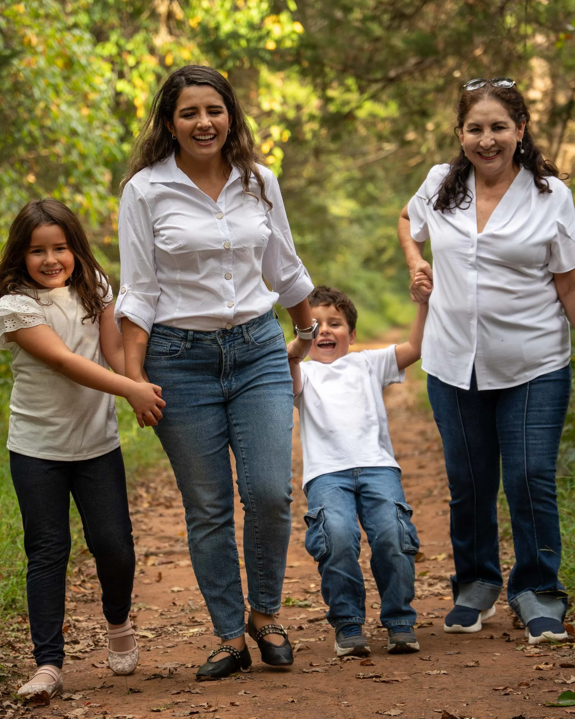 A mom, grandmother and two kids walk toward the camera on a dirt trail through the woods, laughing, as the young boy leans back between his mom and grandmother, holding both their hands.