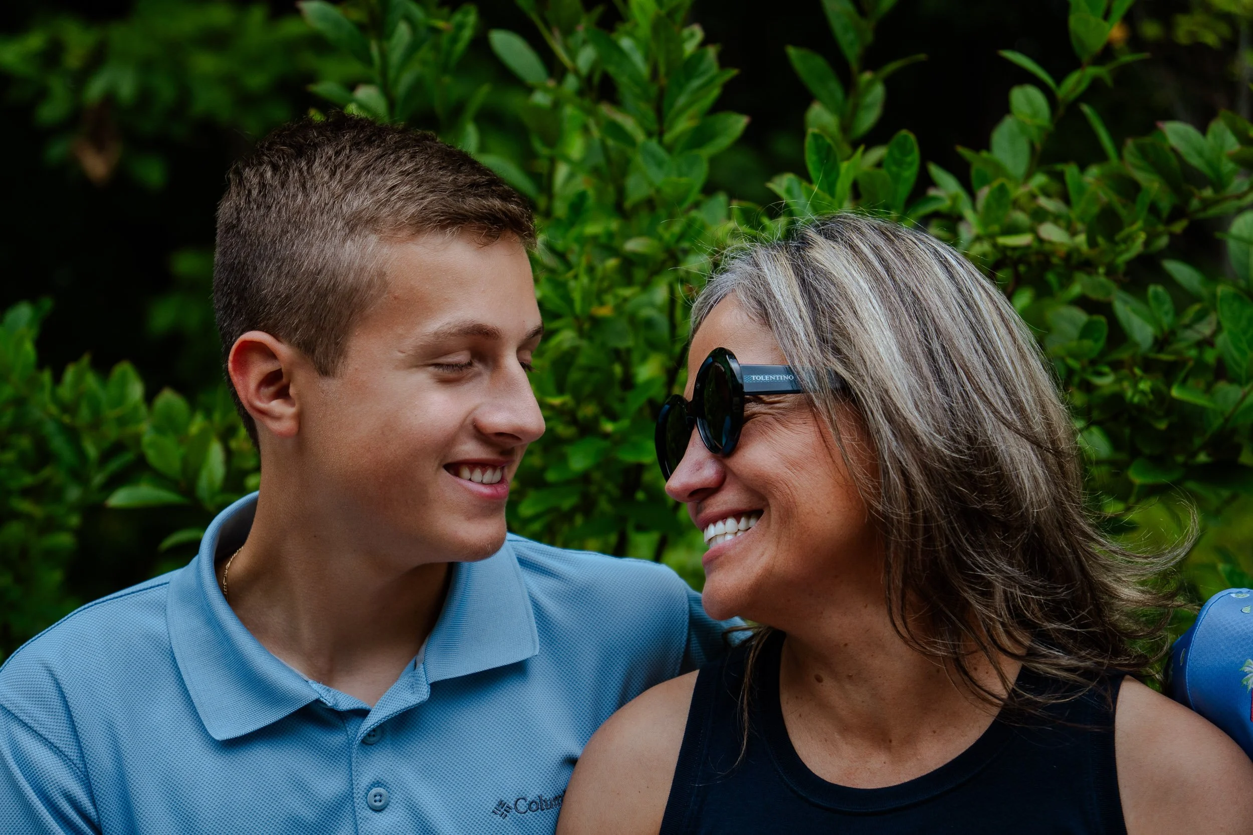 A young man and an older woman smiling and looking at each other, outdoors with lush green foliage in the background. The woman is wearing sunglasses and a black tank top, while the man is wearing a light blue collared shirt.