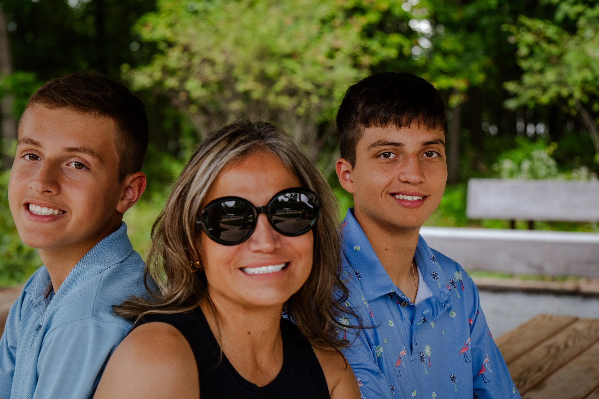 A group of people sitting on a park bench, including a woman with sunglasses and three young men, all smiling and enjoying the outdoors with trees in the background.