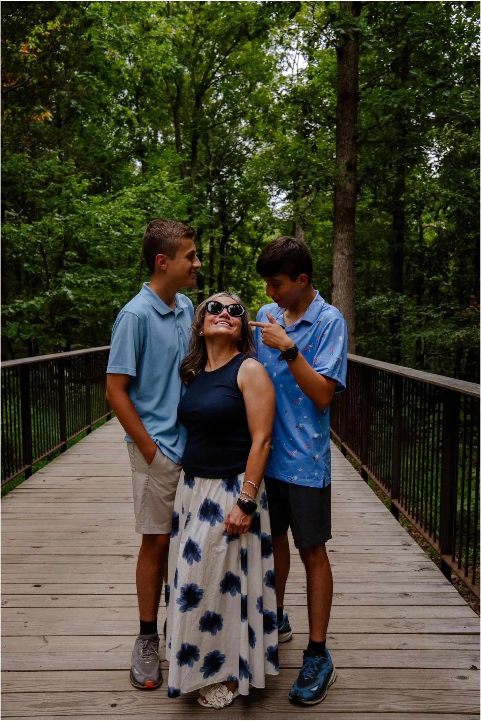 Three people stand on a wooden bridge surrounded by trees, smiling and enjoying each other's company.